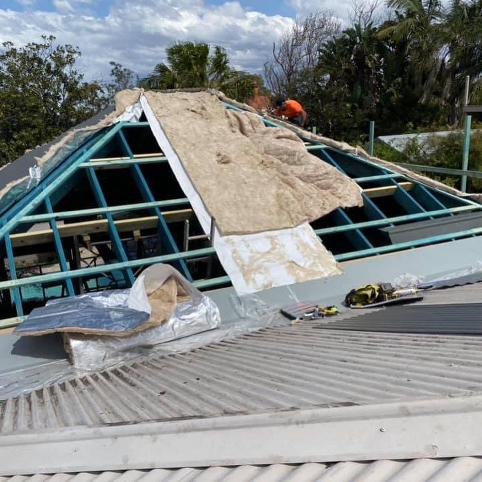 Collapsed roof under repair with workers on a damaged building frame, exposed blue beams, and tarp covering debris