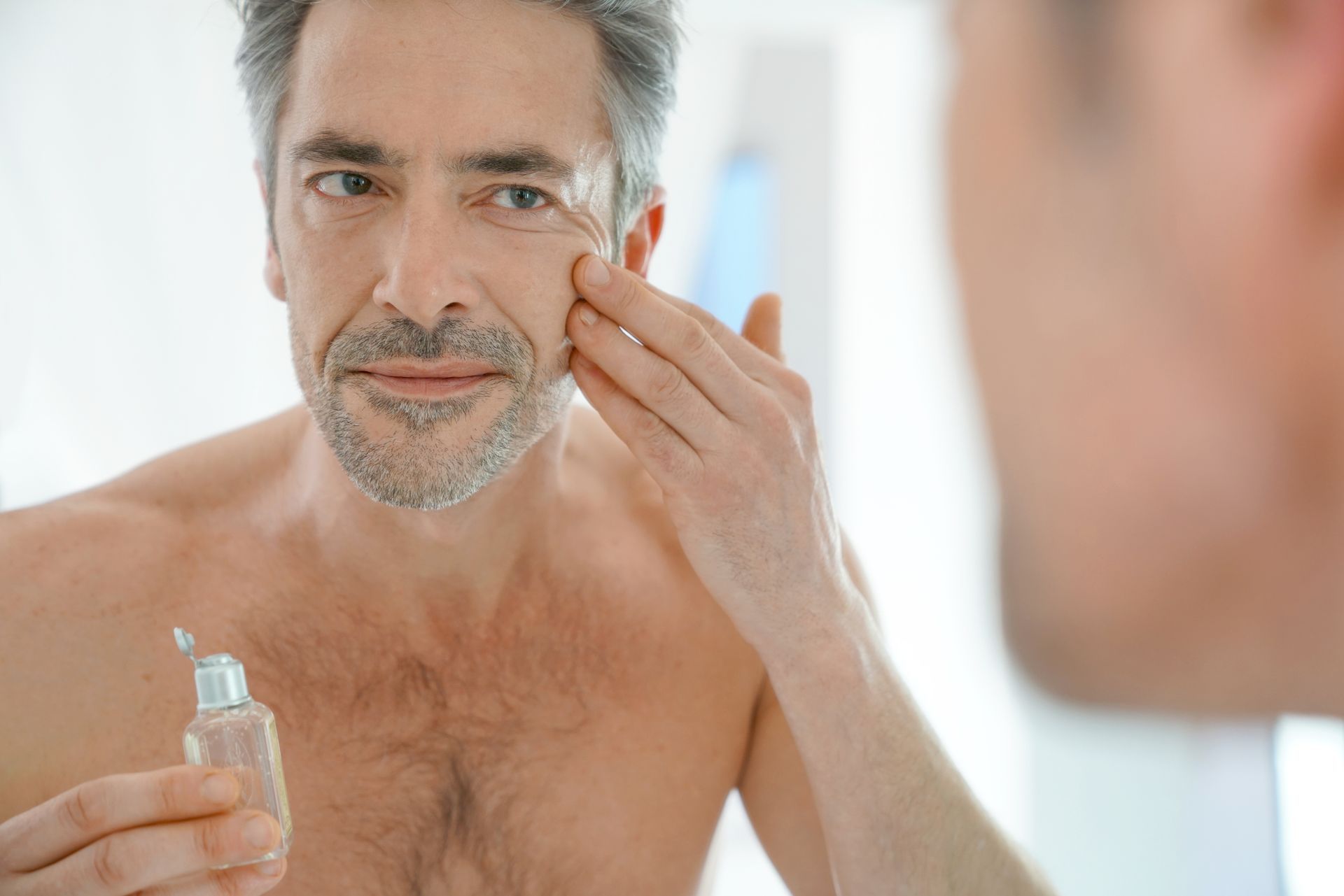 Ein Mann ohne Hemd trägt vor einem Spiegel Lotion auf sein Gesicht auf.