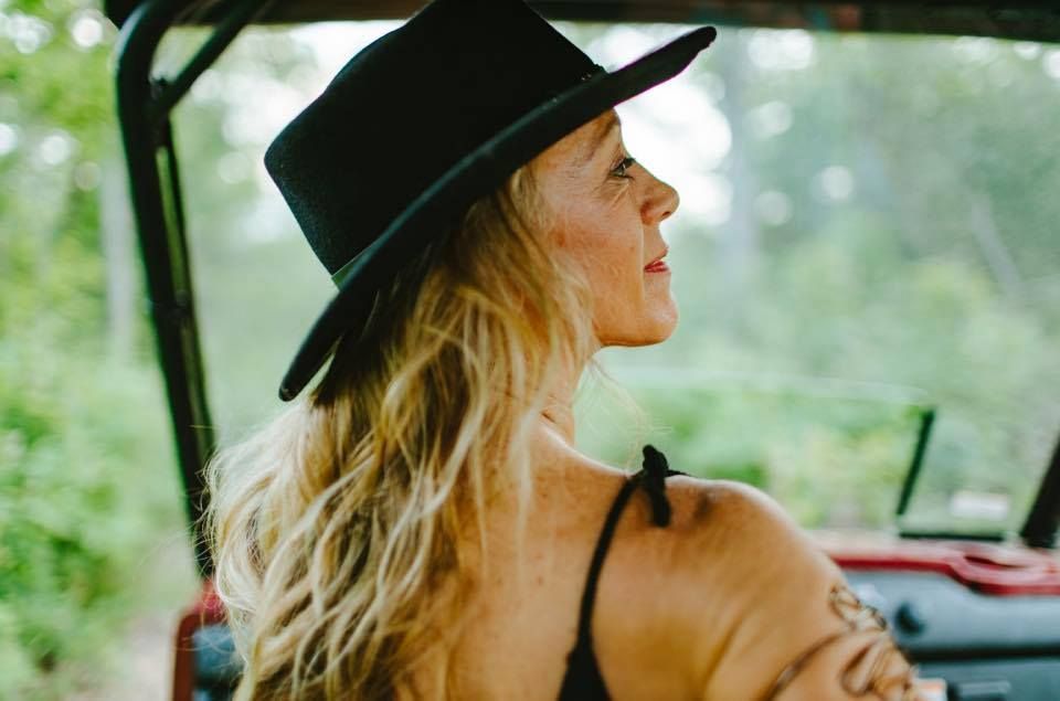 Franny Tacy driving an off-road vehicle, looking to the side. Blonde hair, trees in background.