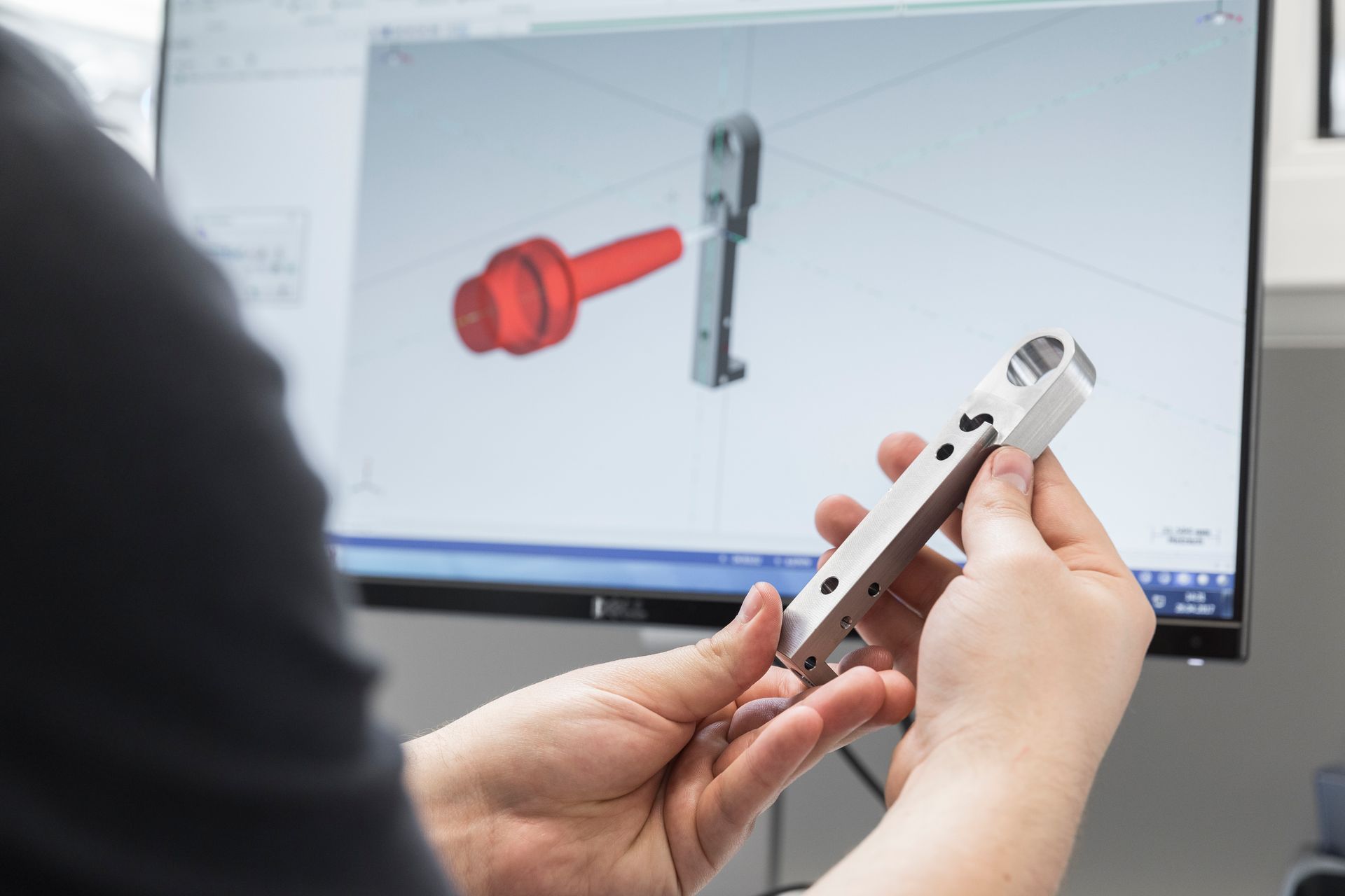 Eine Person untersucht ein Metallteil und vergleicht es mit einem 3D-Modell auf einem Computerbildschirm.