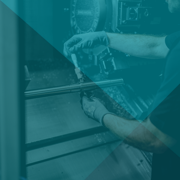 Person mit Handschuhen arbeitet in einer Fabrikumgebung mit Maschinen an einer Metallstange.