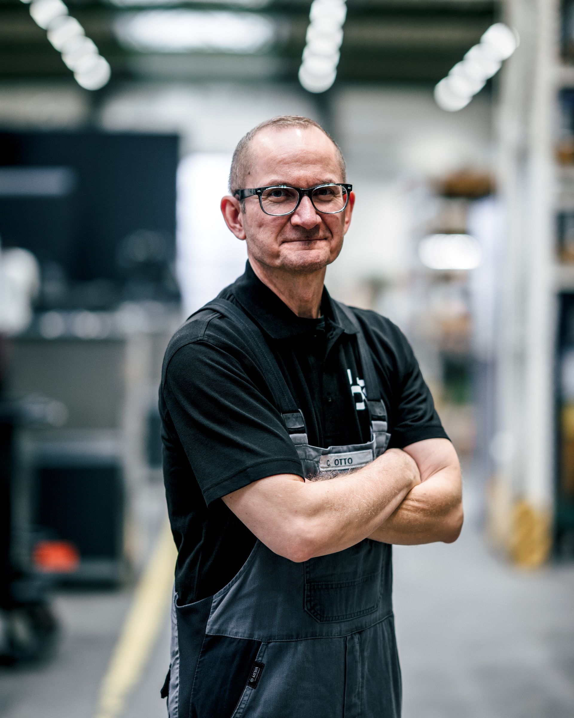 Mann in einer Werkstatt, Brille, Arme verschränkt. Er trägt ein schwarzes Hemd und einen Overall. Industrieumgebung.