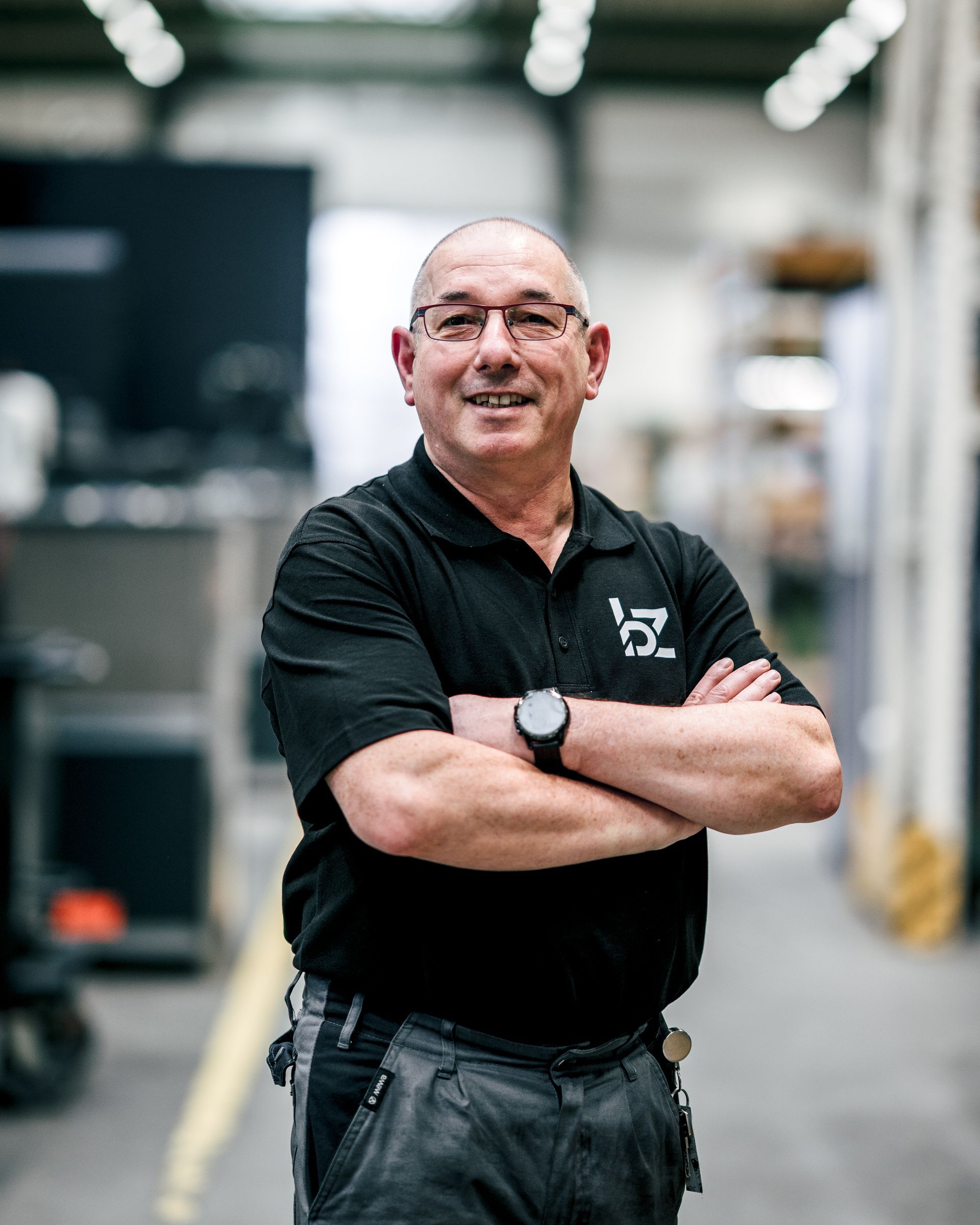 Mann in schwarzem Poloshirt mit verschränkten Armen in einer Fabrikumgebung.