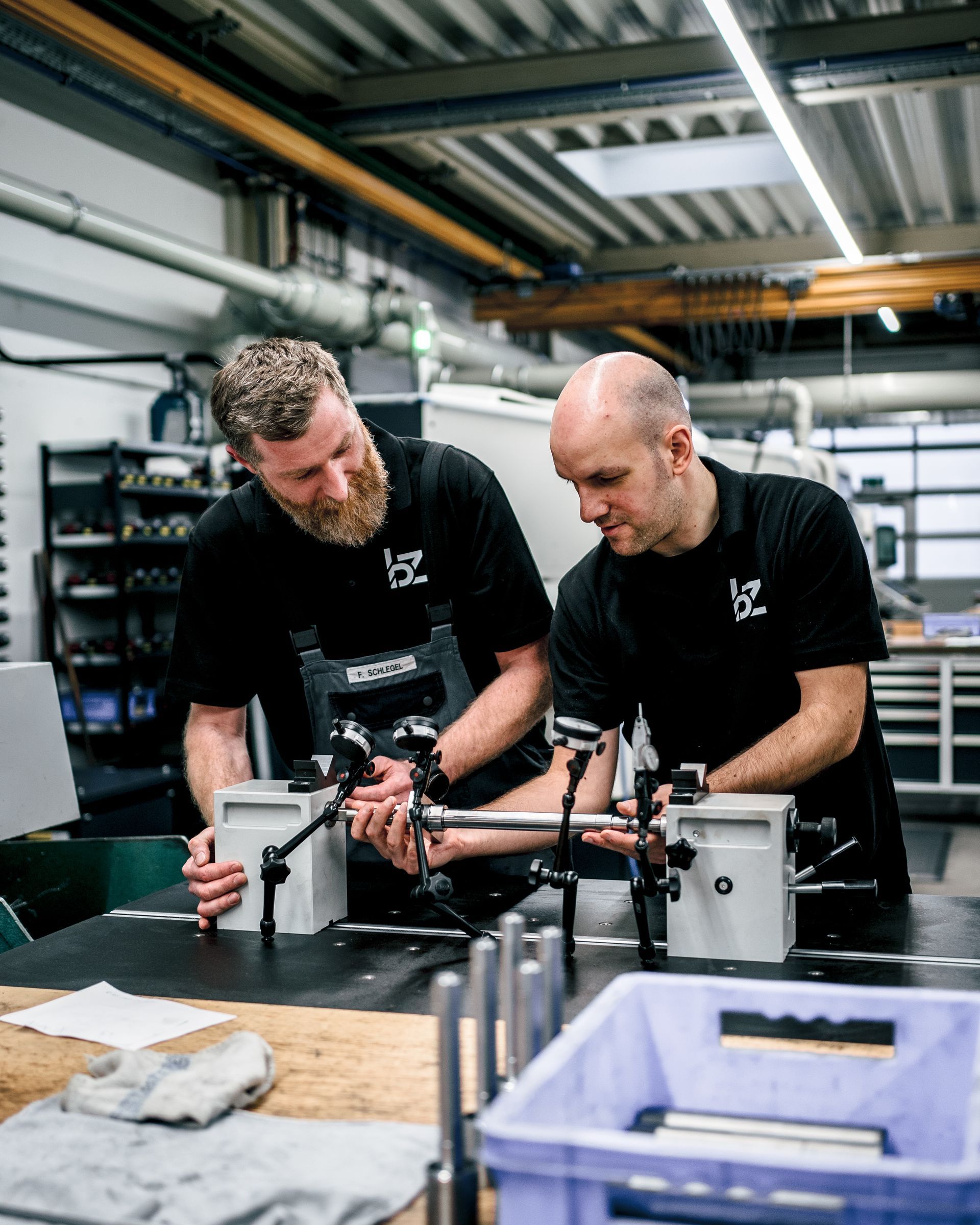 Zwei Männer in schwarzen Hemden untersuchen Metallteile an einer Werkbank in einer Werkstatt.