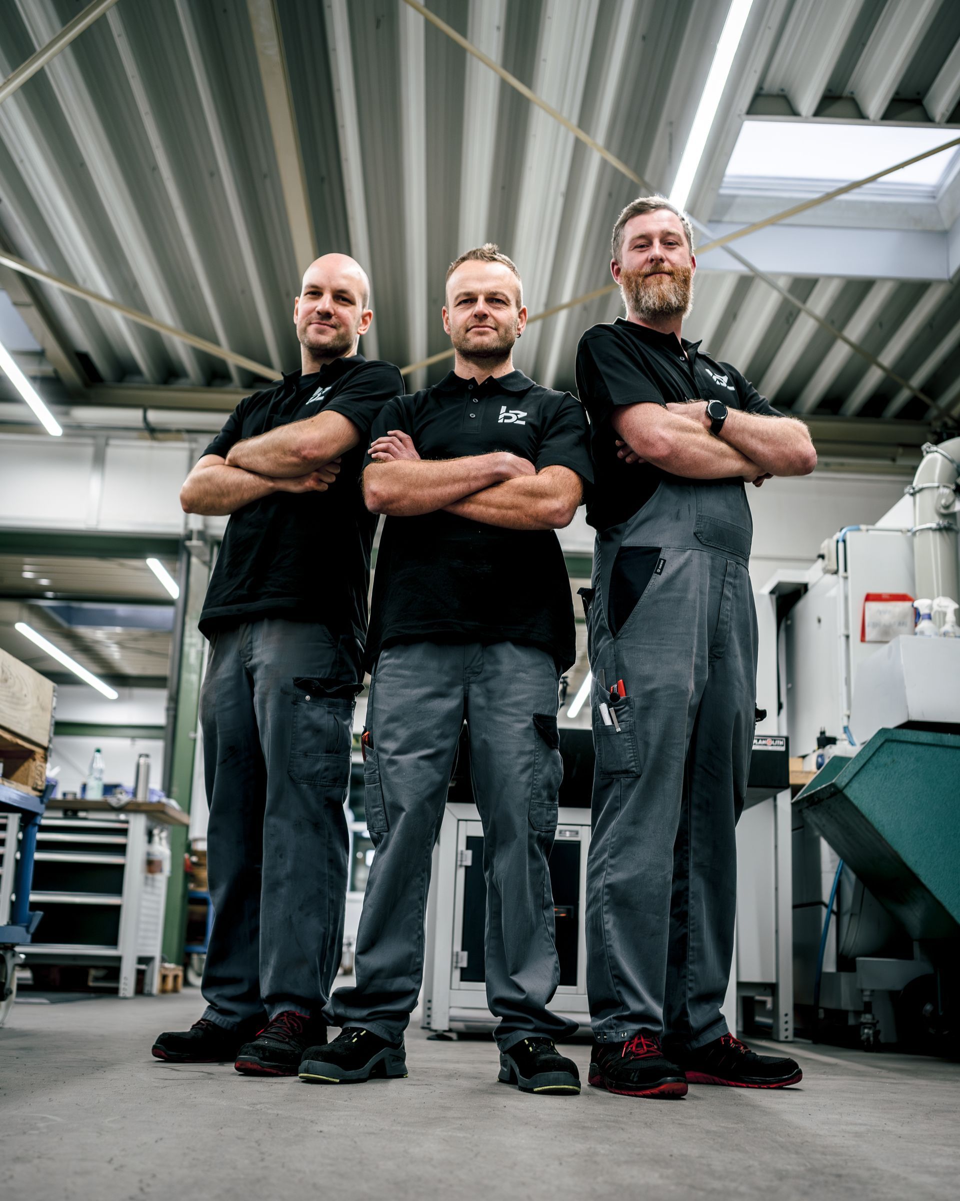 Drei Personen in Arbeitskleidung stehen mit verschränkten Armen in einer Werkstatt.