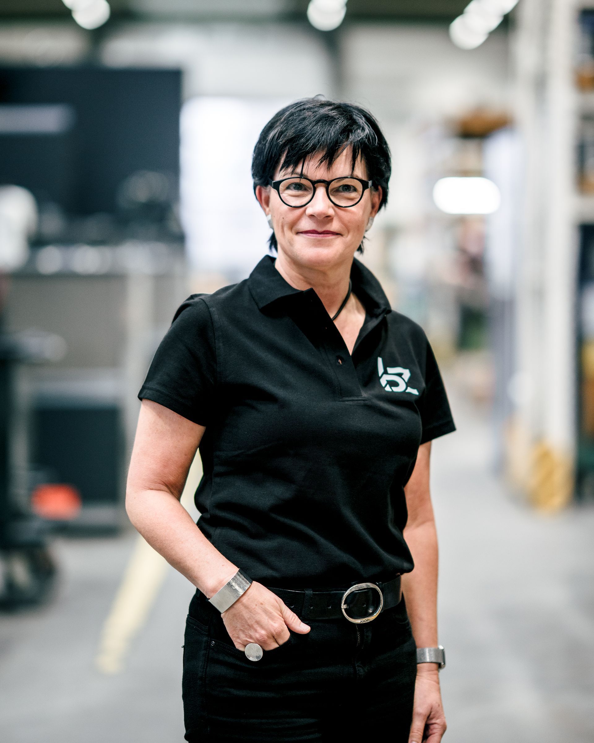 Frau in schwarzem Poloshirt und Brille, lächelnd, in einer Lagerhalle.
