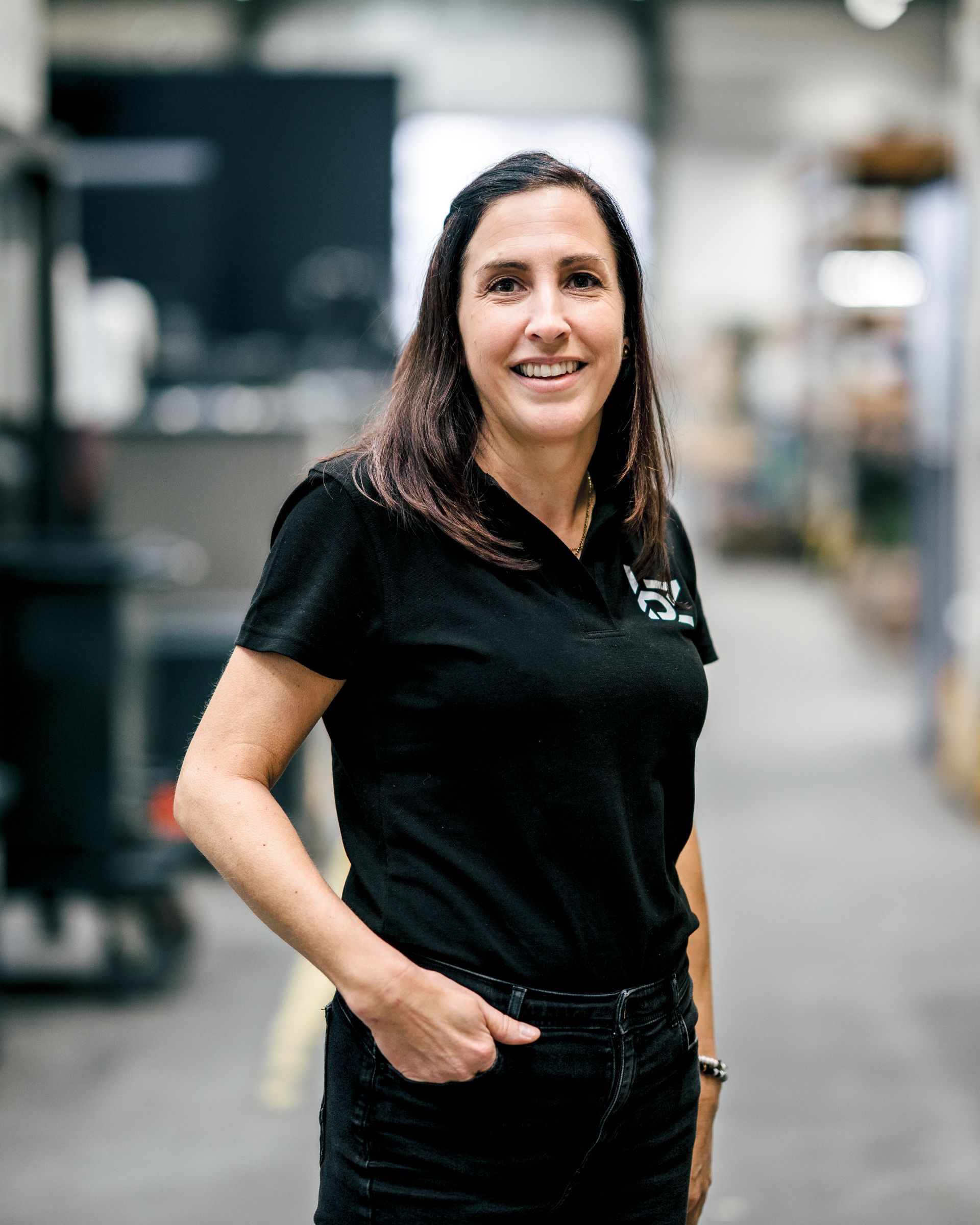 Frau in schwarzem Hemd und Jeans lächelt in einer Werkstattumgebung, die Hand in der Tasche.
