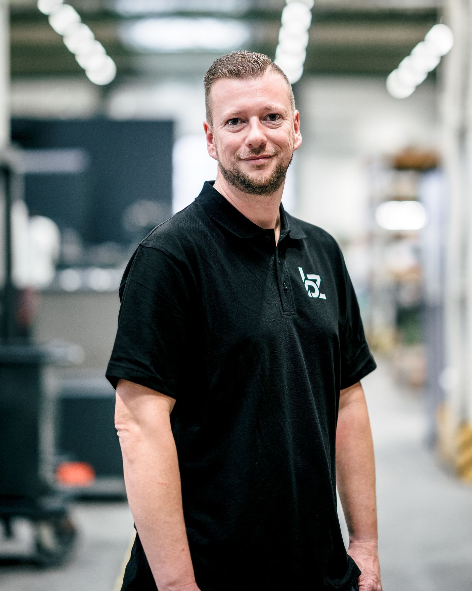 Ein Mann in einem schwarzen Poloshirt lächelt in einer Fabrikhalle; auf dem Shirt ist ein Logo zu sehen.