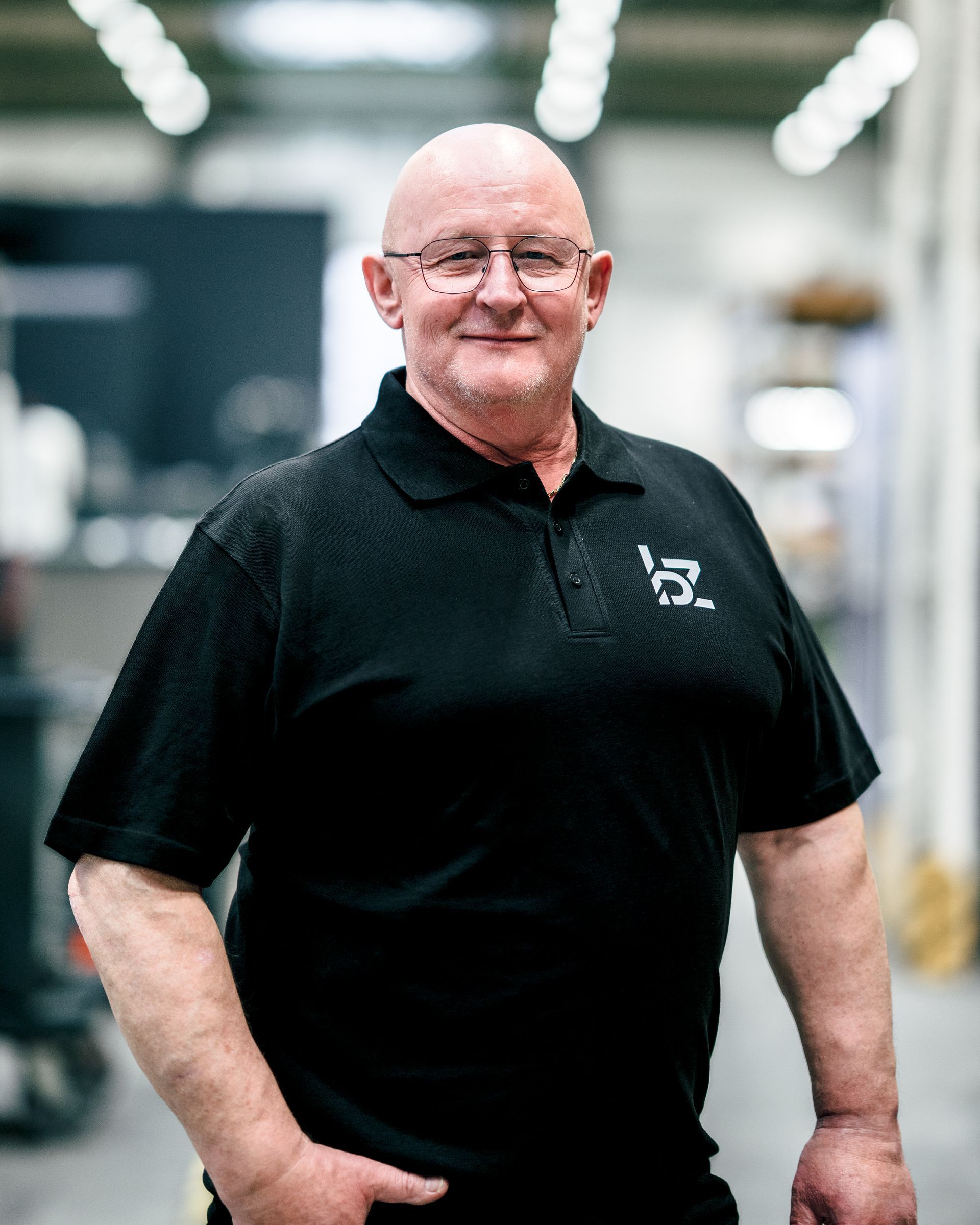 Ein Mann mit Brille und schwarzem Poloshirt steht in einer Fabrikhalle.