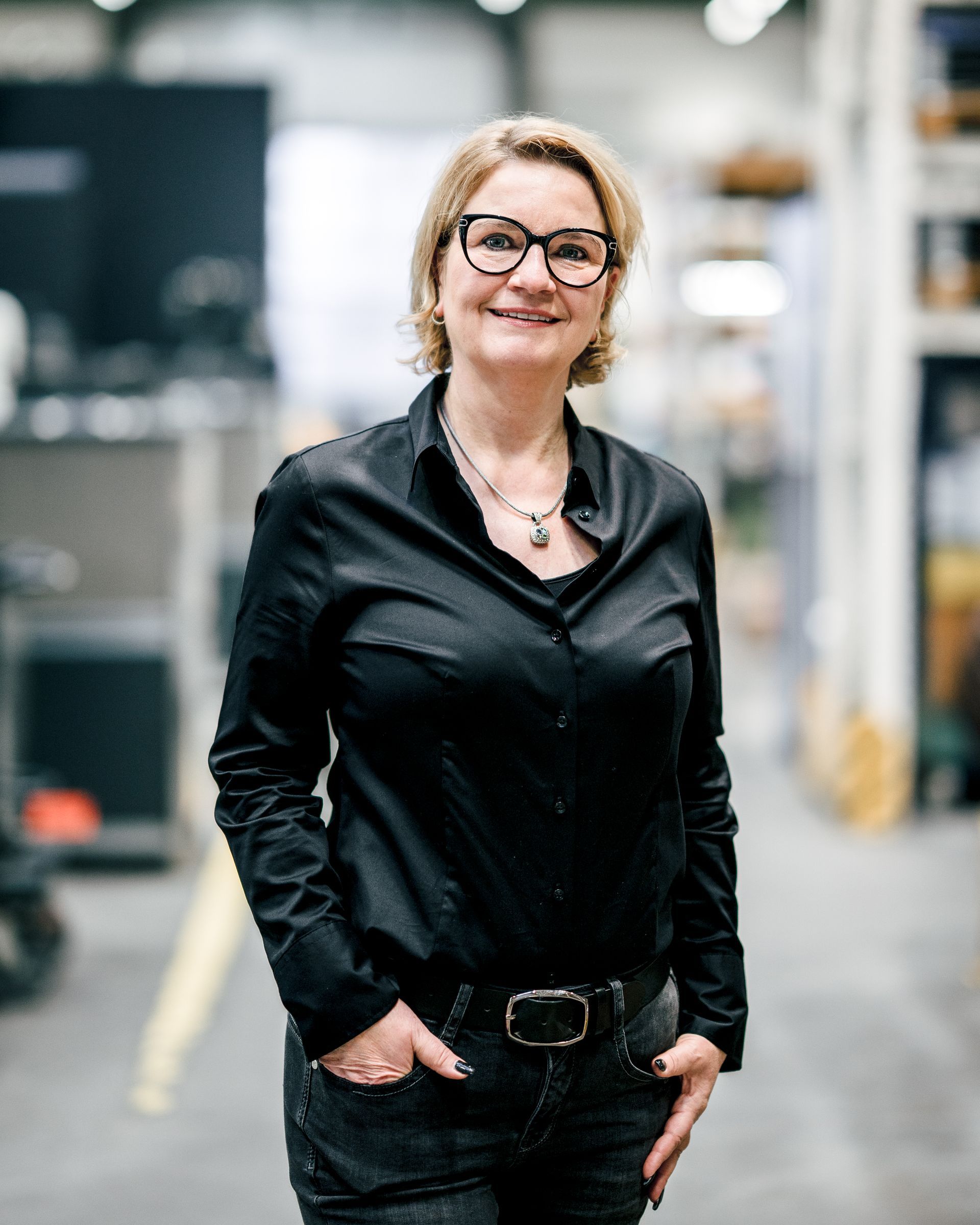 Eine Frau in schwarzem Hemd und Brille lächelt, steht mit den Händen in den Taschen in einer Lagerhalle.