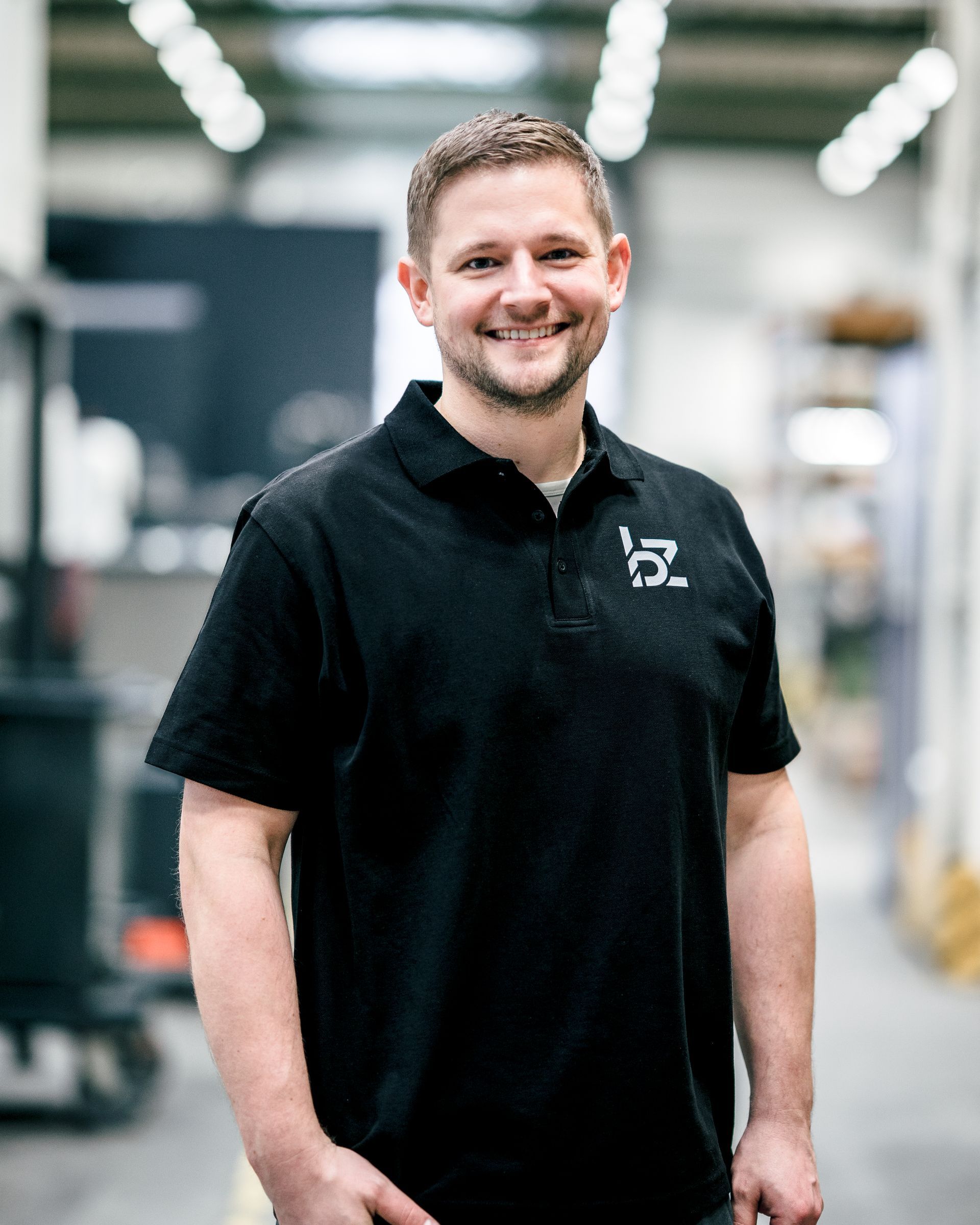 Mann in schwarzem Poloshirt lächelt in Industrieumgebung.