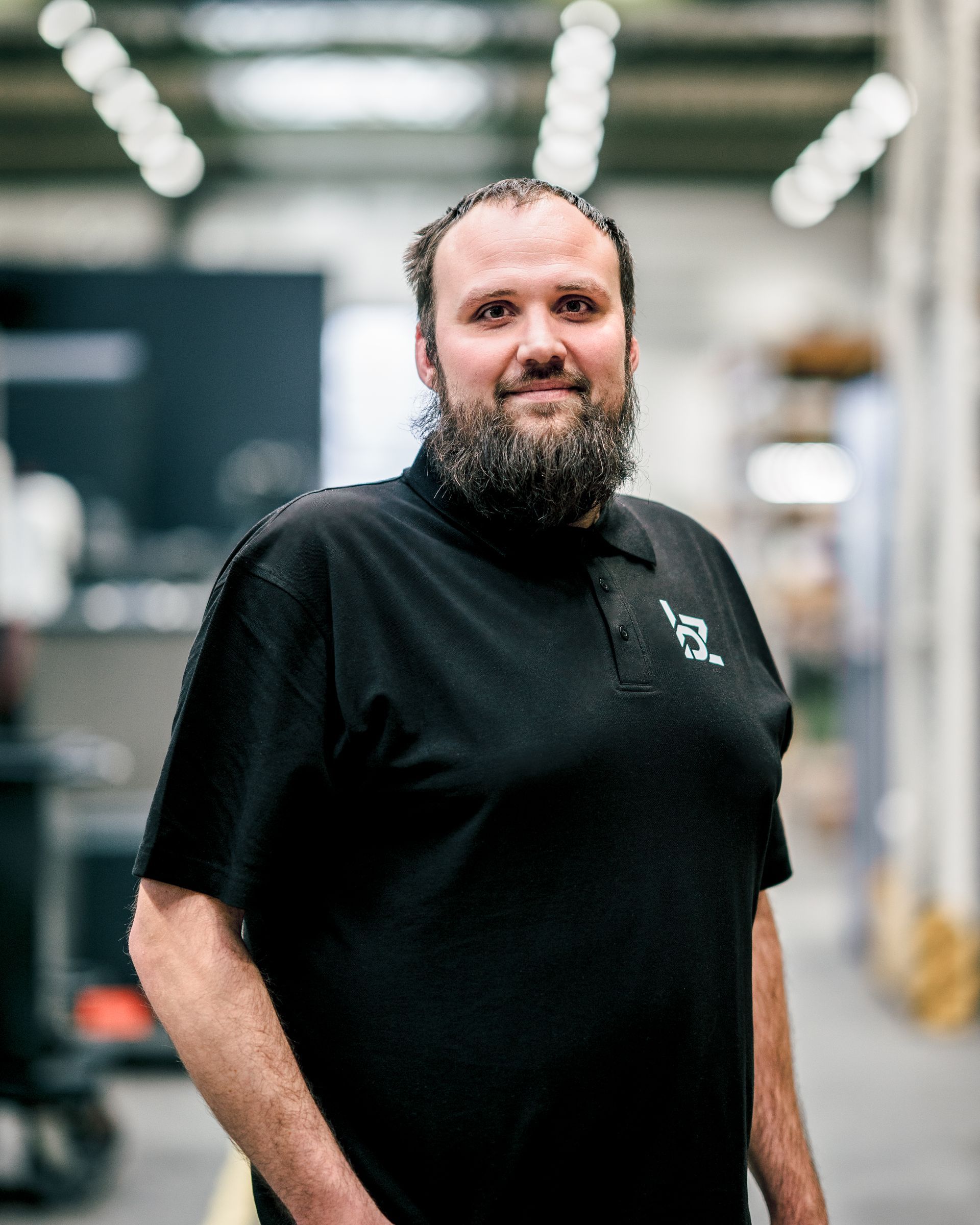 Mann mit Bart und schwarzem Hemd steht in einem Fabrikgang.