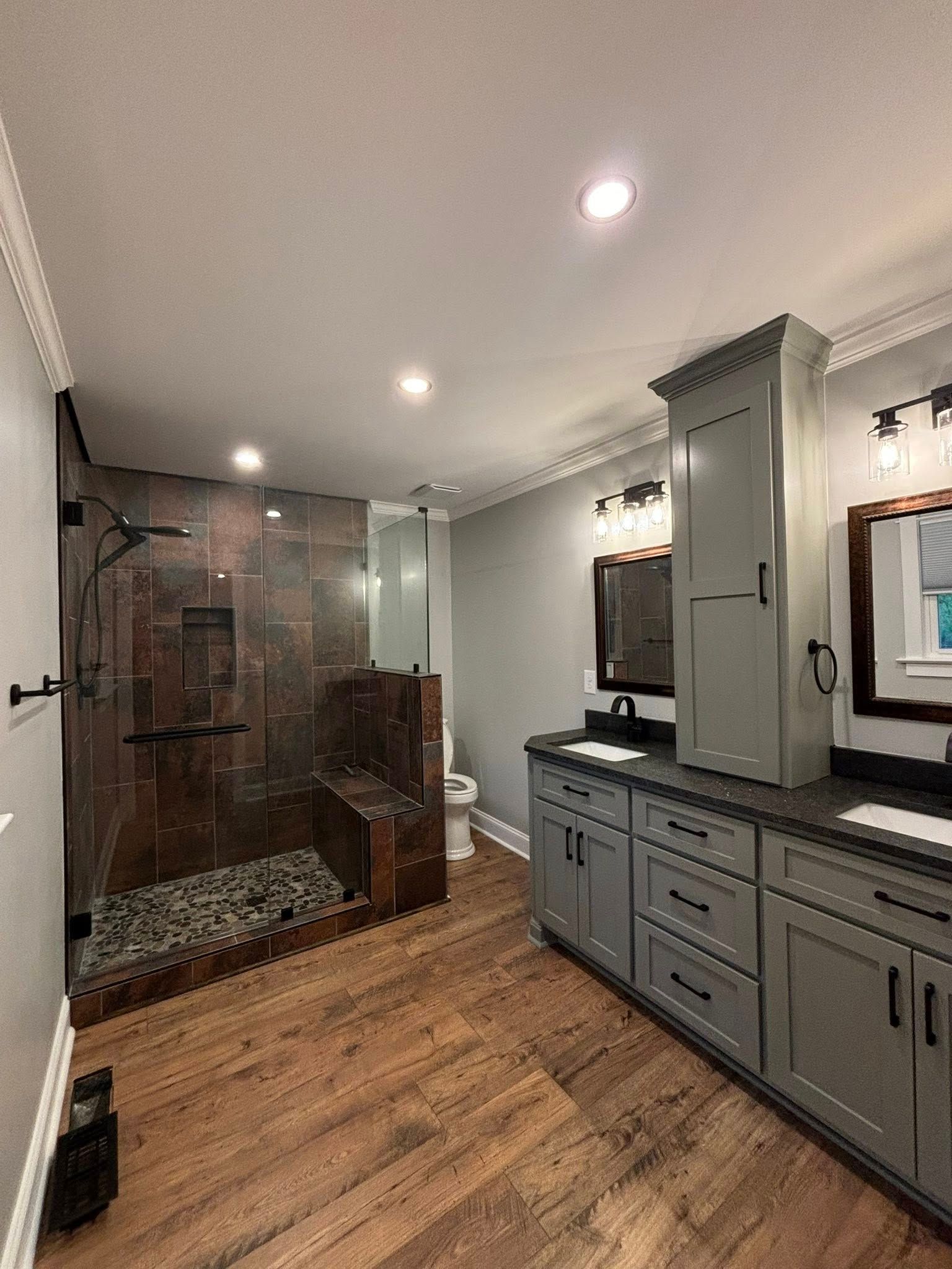A bathroom with hardwood floors , gray cabinets , a toilet , and a walk in shower.