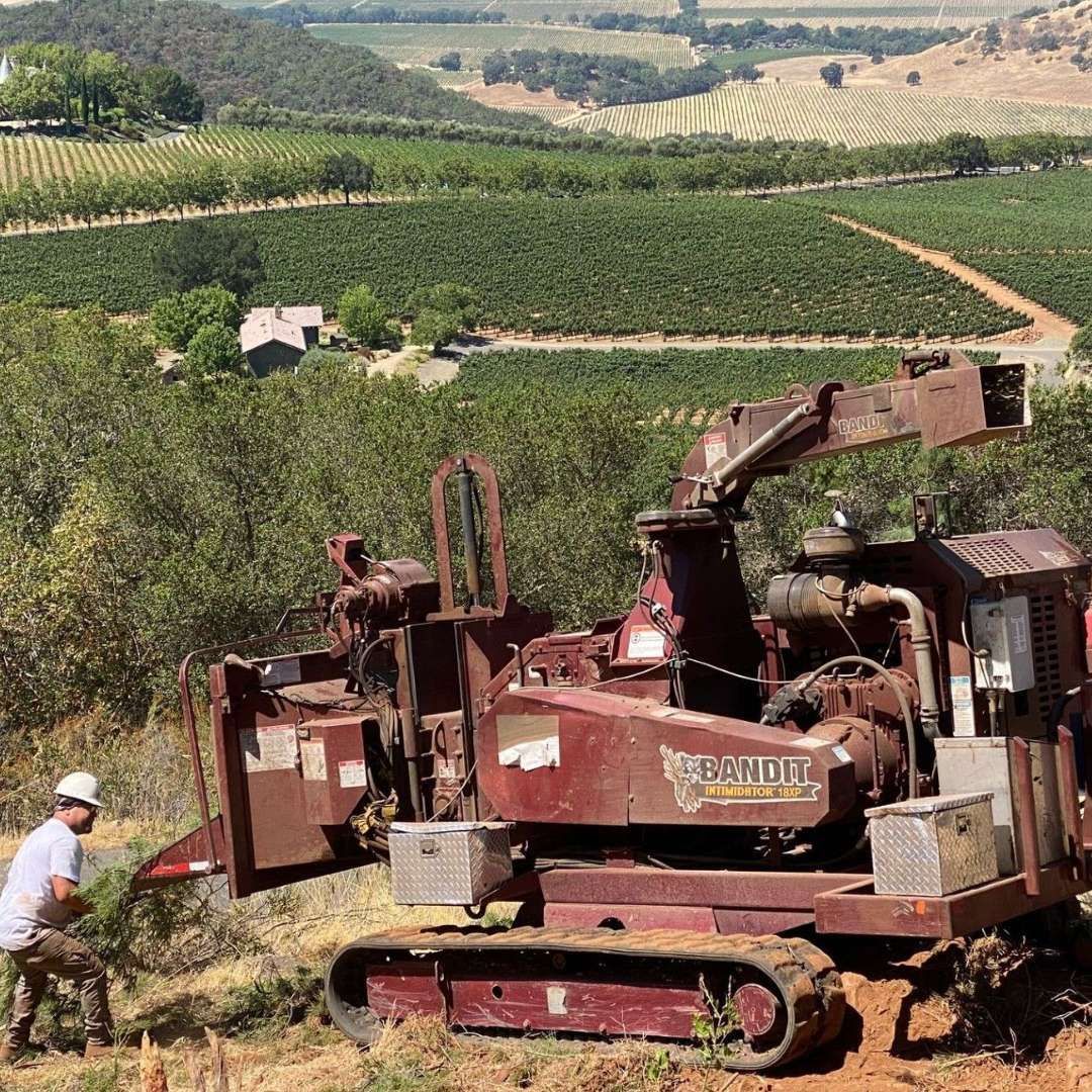 Man operating a red grinder on a hillside overlooking vineyards and a valley. Man operating a red grinder on a hillside overlooking vineyards and a valley.
