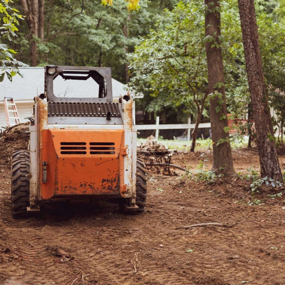 An orange skid steer clears dirt in a yard with trees, near a house with a white fence. An orange skid steer clears dirt in a yard with trees, near a house with a white fence.