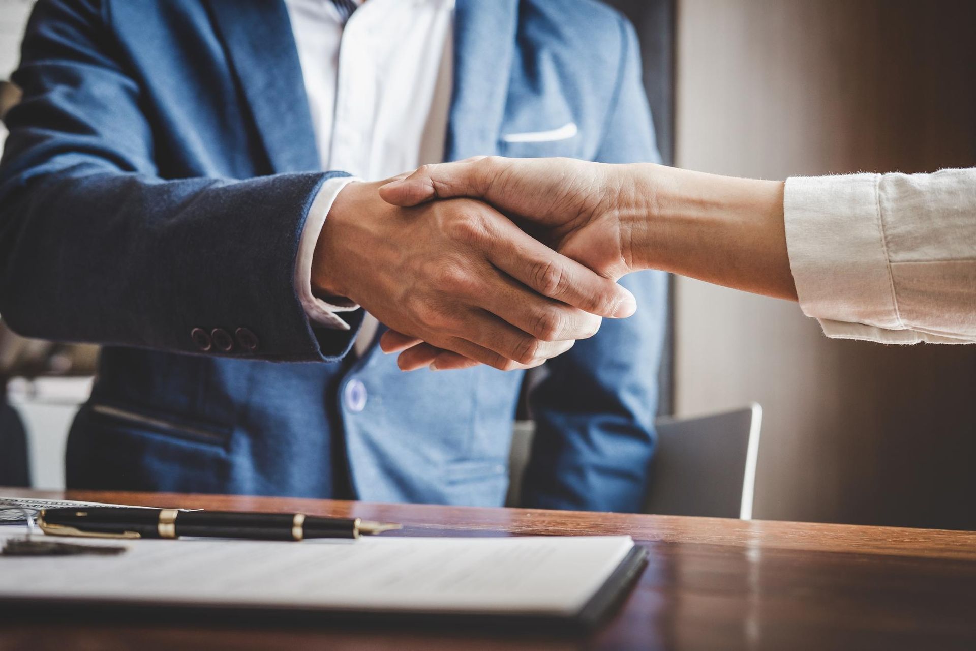 Man in blue suit shakes hands with person in white shirt, over a desk with a contract.