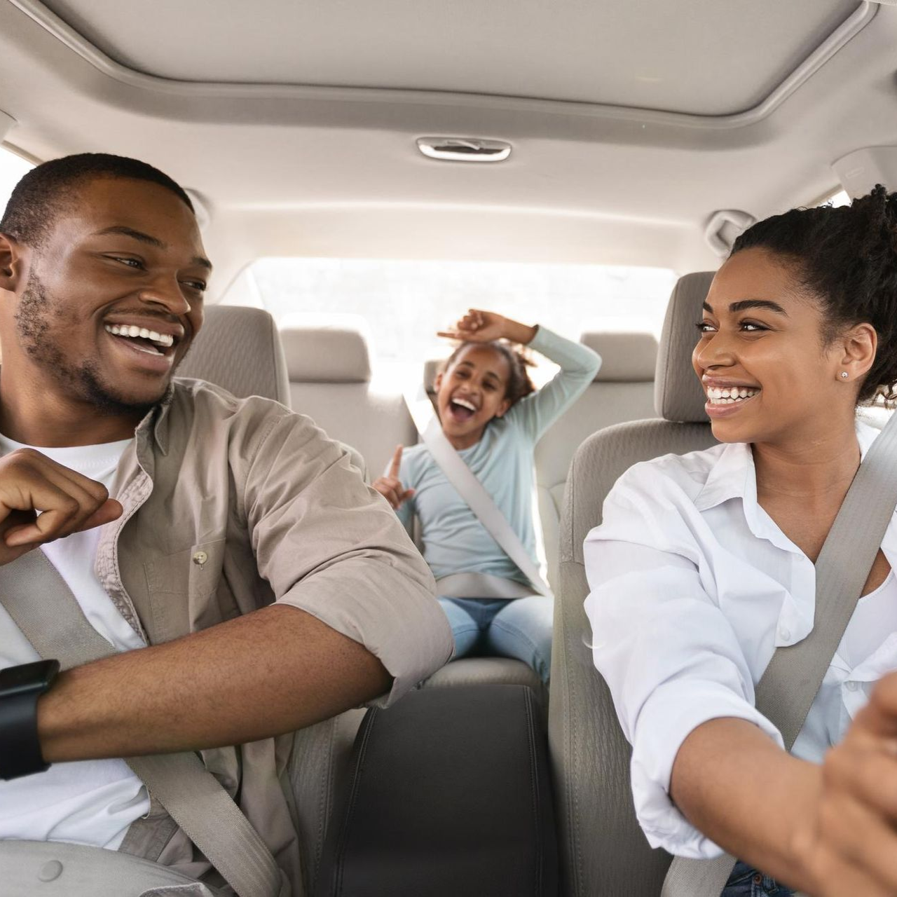Family in a car, smiling and laughing. Child in back, hands up. Inside of car, sunny.