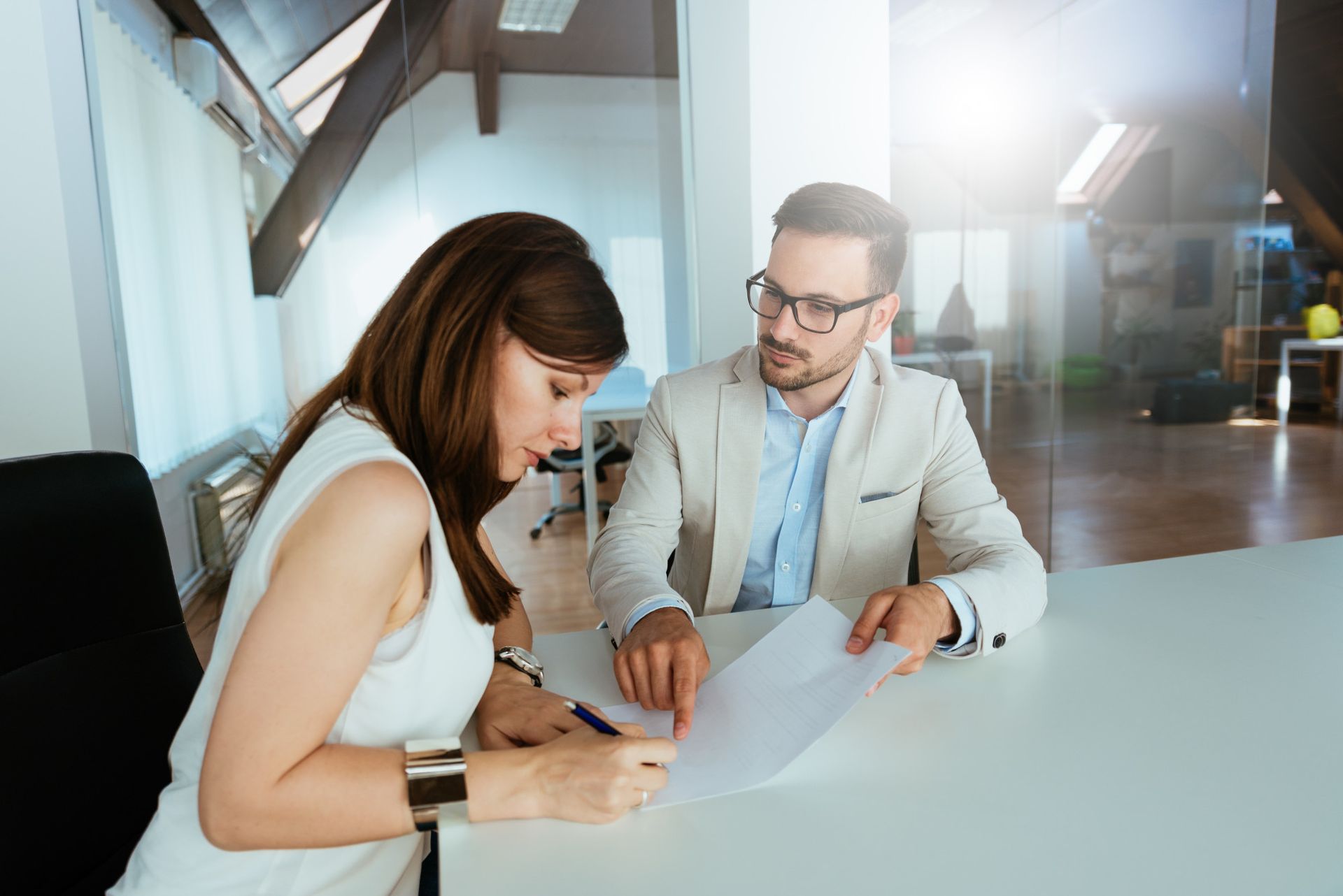 Woman signing document at desk, advised by a man in a light blazer. Bright office setting.
