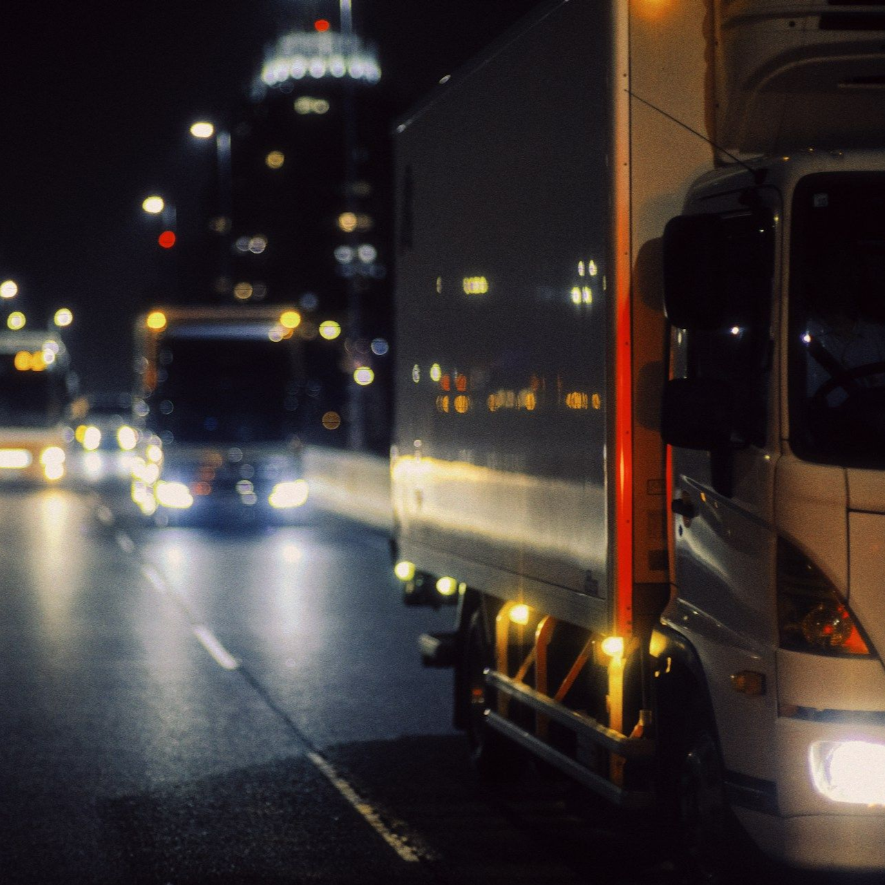 Truck driving at night on a city street, headlights illuminating wet asphalt, with blurred background of city lights.