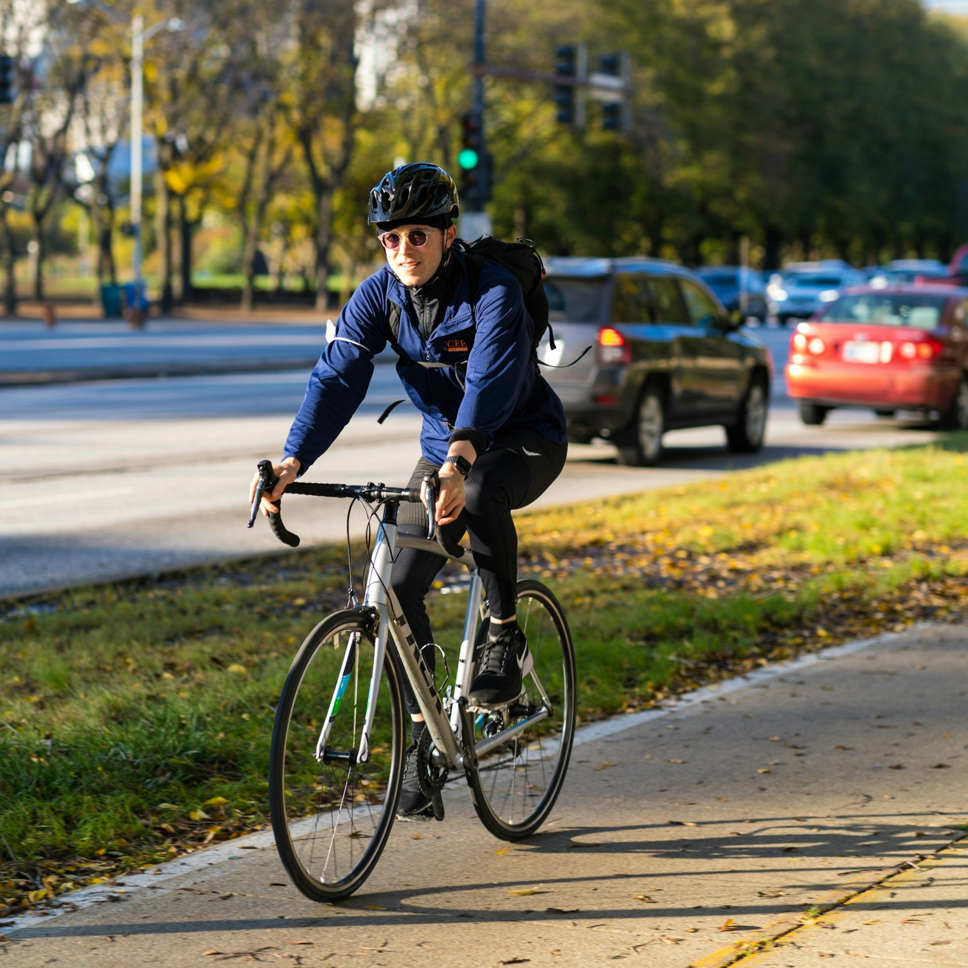 Person in helmet and jacket riding bicycle on a path next to a road with cars in the daytime.