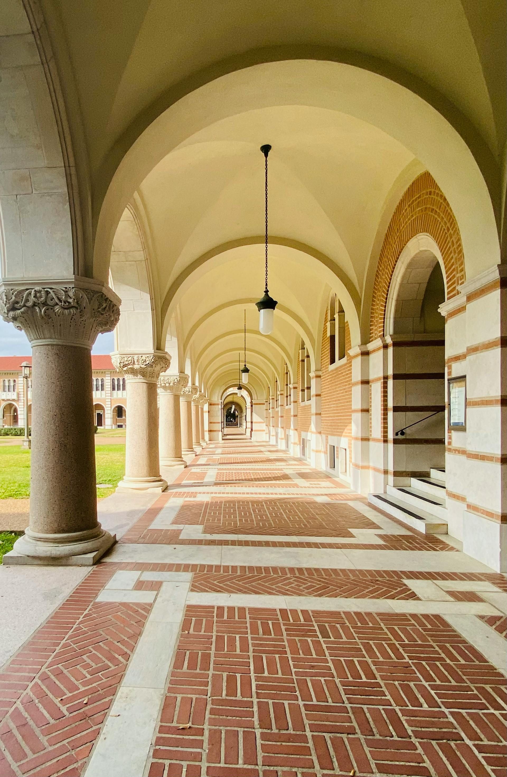 Arched walkway with brick floor, columns, and a distant view; sunlight illuminates the corridor.
