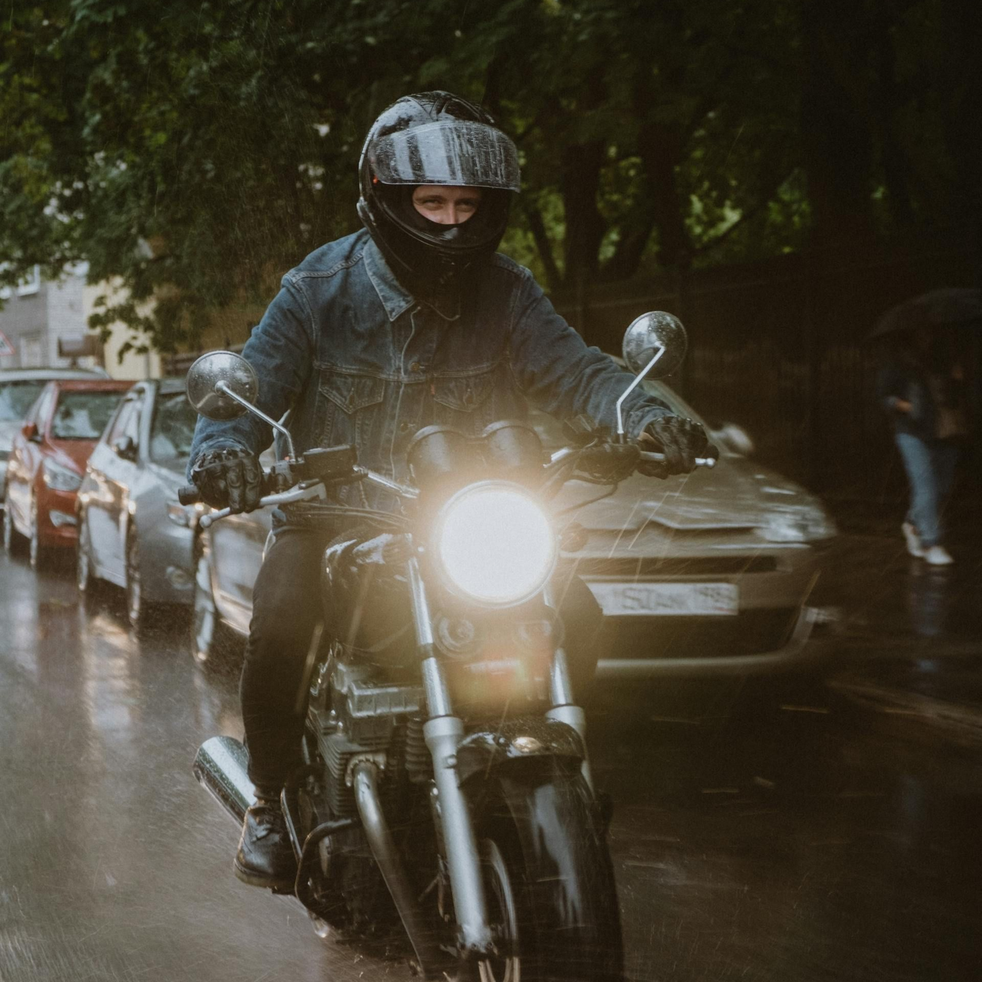 Person on motorcycle in the rain, wearing a helmet and black face covering, traveling on a wet road near parked cars.