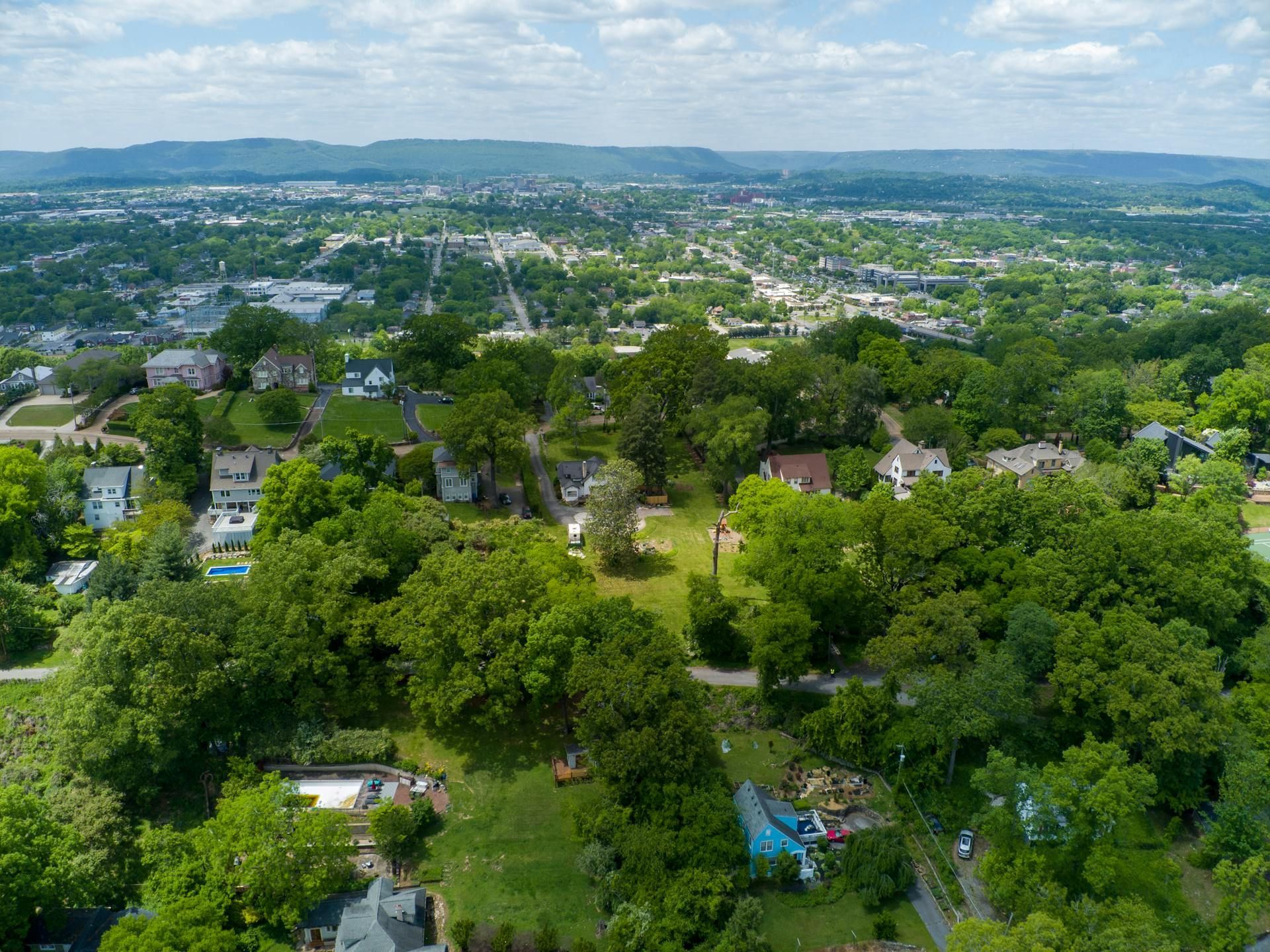 Overhead view of a town with houses, trees, and a distant cityscape under a bright blue sky.
