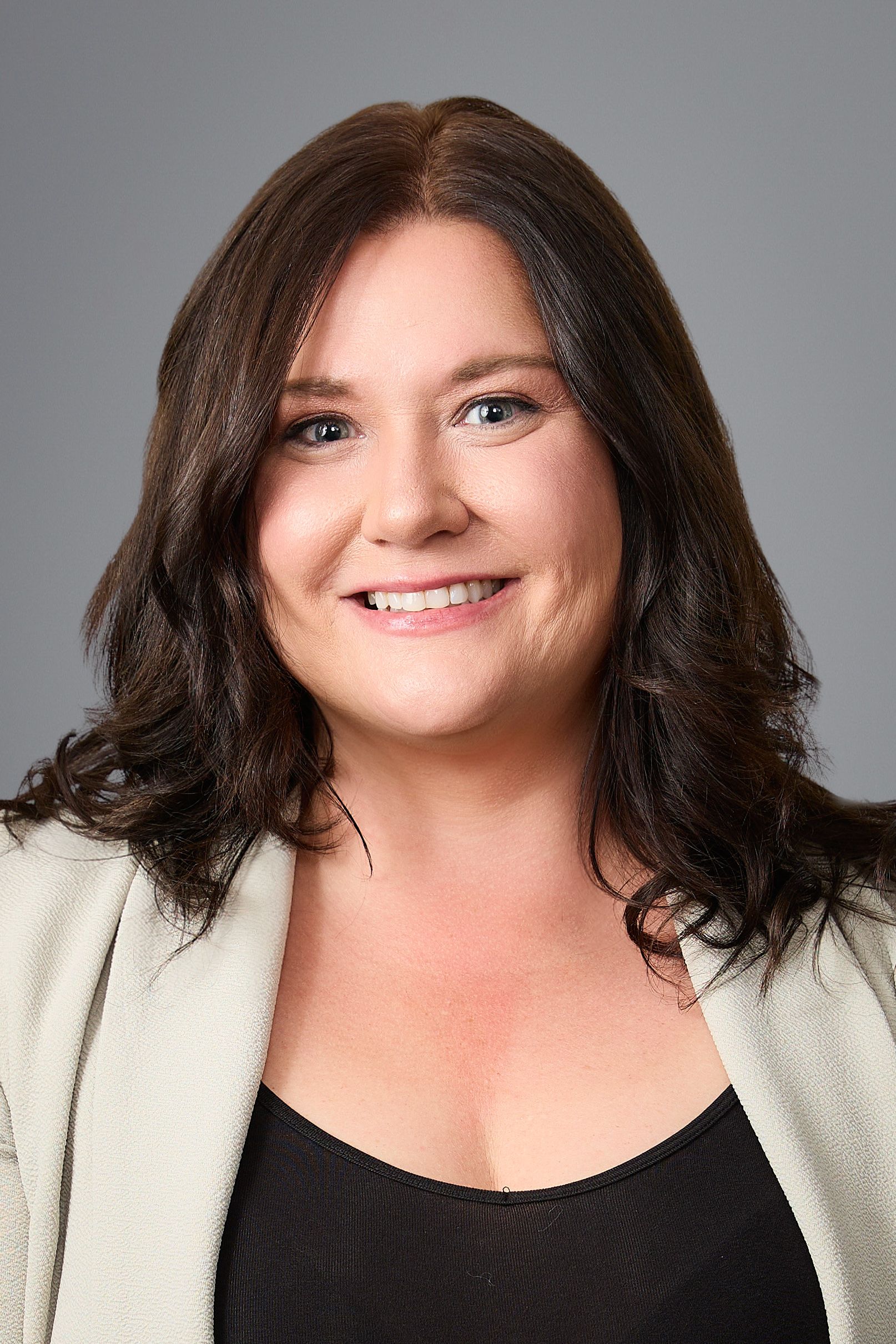 Woman with dark hair, smiling, in a blazer against a grey background.