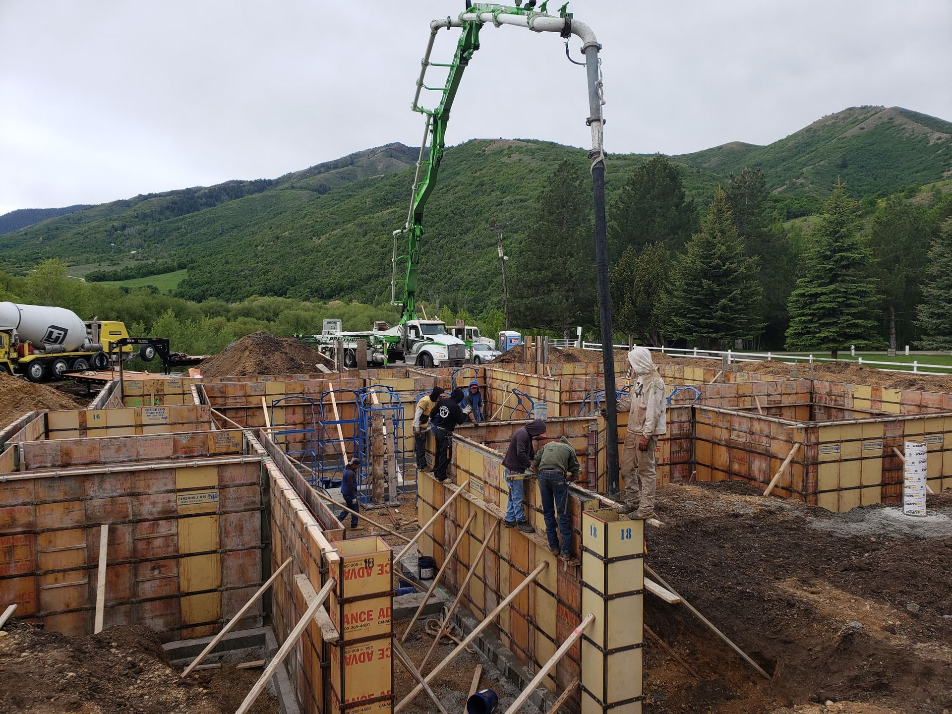Construction site with workers pouring concrete from a boom pump into foundation forms, backed by green hills