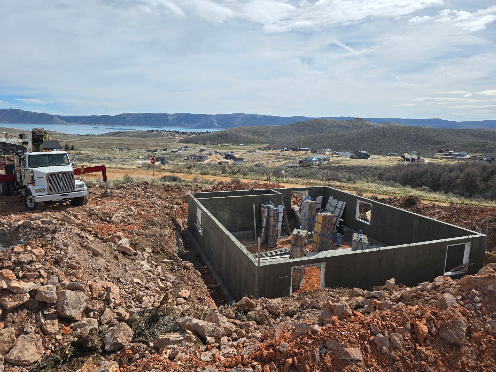 Construction site with foundation walls, excavator, and white truck on rocky terrain overlooking distant hills