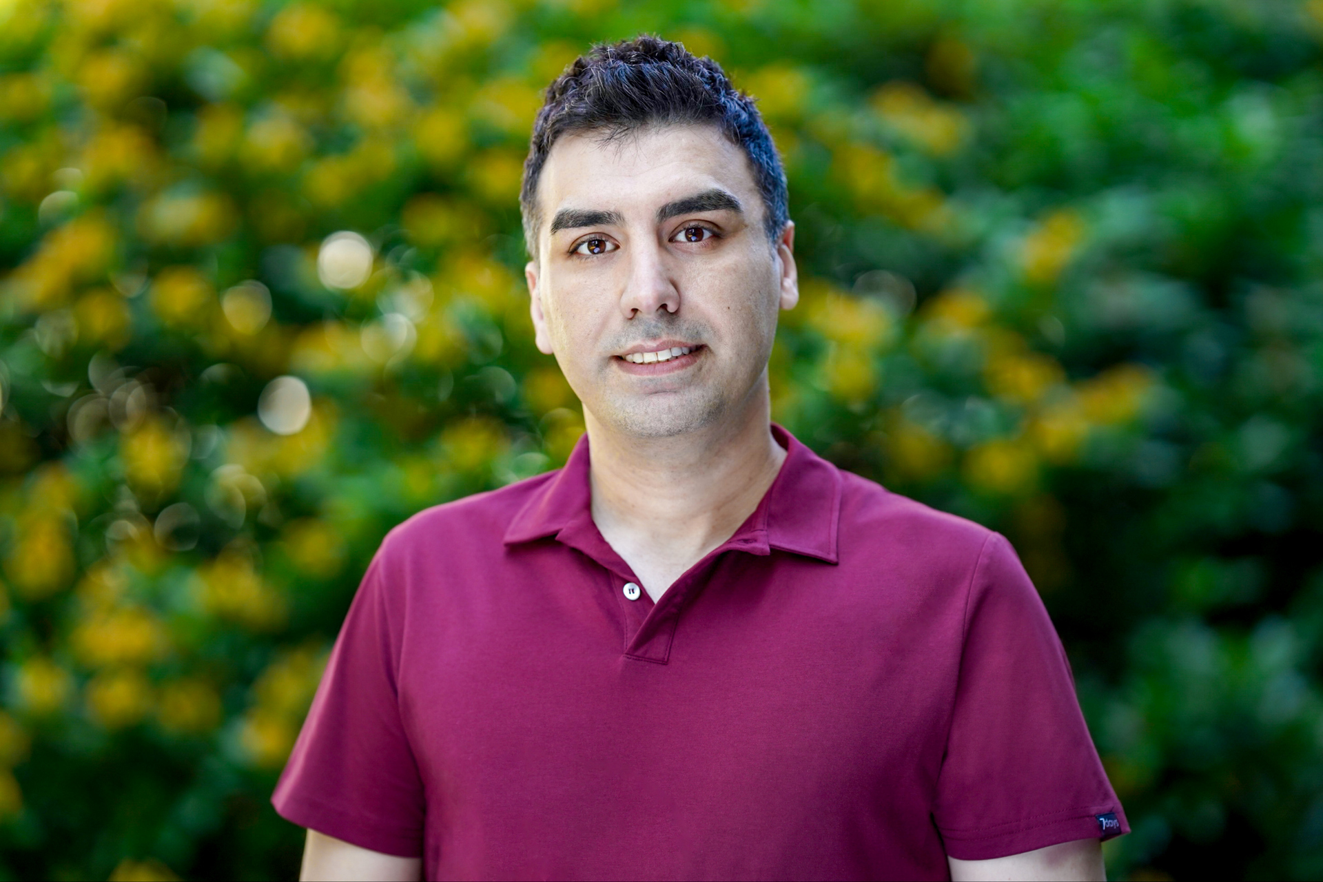 En mann i en rødbrun skjorte står foran en busk med gule blomster.