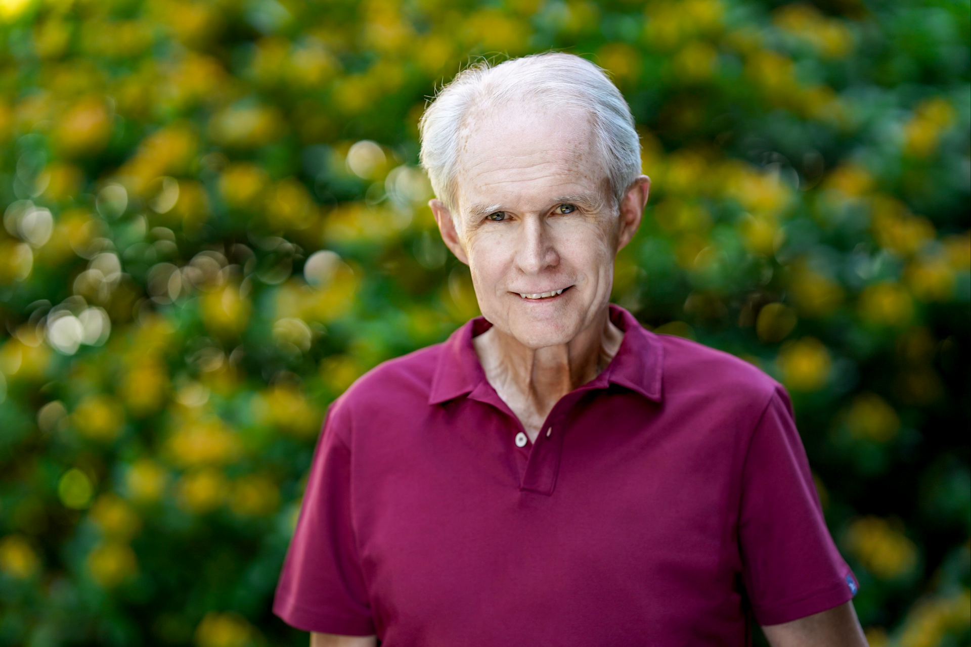En eldre mann i rød skjorte står foran en busk med gule blomster.