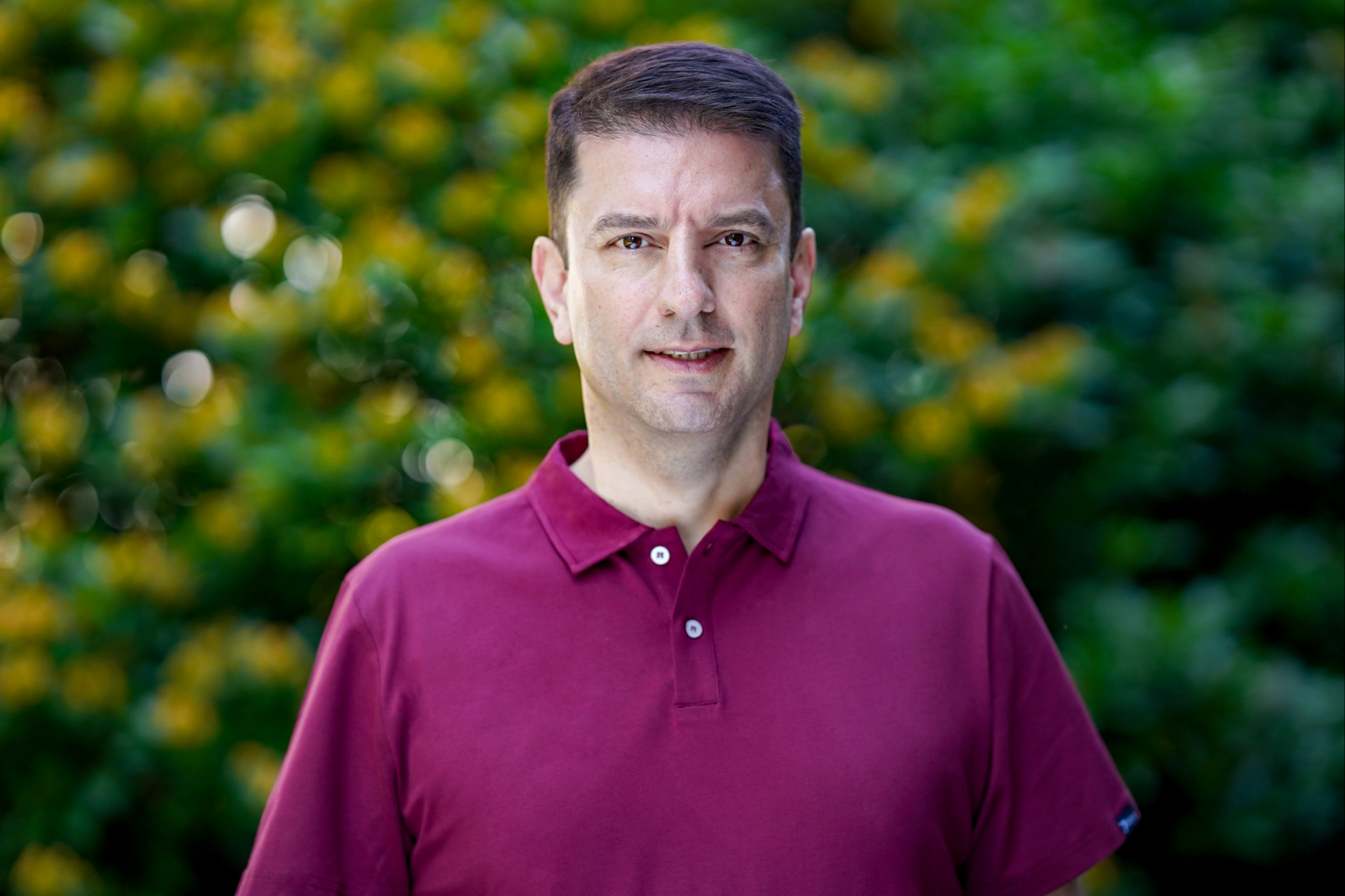 En mann i en rødbrun poloskjorte står foran en busk med gule blomster.