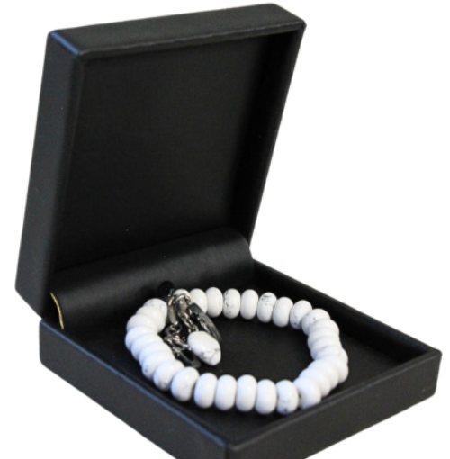 Witte kralenarmband met schedelbedel in een zwart sieradendoosje op een houten oppervlak.