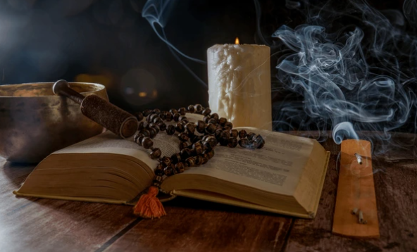Een open boek, een rozenkrans, brandende kaarsen, rook en een kom op een houten oppervlak.