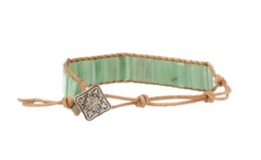 Armband van groene rechthoekige kralen, omwikkeld met bruin leer, met zilveren bedel, rustend op een witte steen.