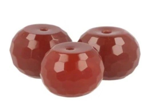 Drie gefacetteerde, roodbruine carneoolkralen op een witte selenietkom. Groene bladeren op de achtergrond.