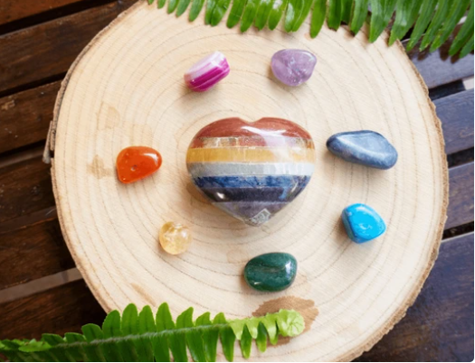 Hartvormige chakrastenen op een houten schijf, omringd door verschillende kleurrijke kristallen en groen.