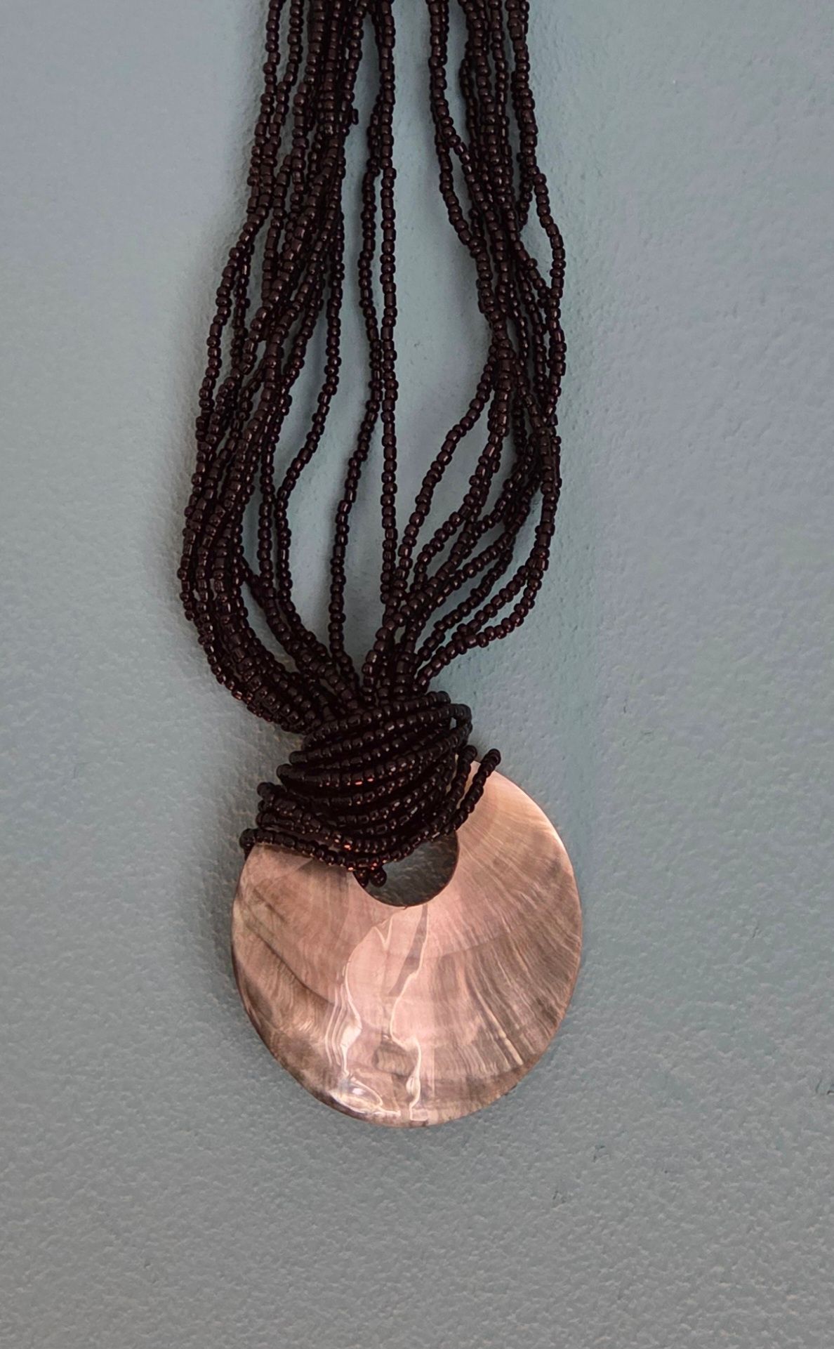 Zwarte kralenketting met een ronde hanger van parelmoer, geplaatst tegen een blauwe achtergrond.