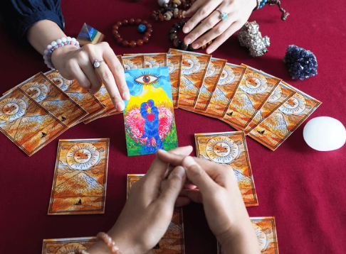 Handen leggen tarotkaarten op een rode tafel. Eén kaart ligt open, met een oog en kleurrijke figuren.