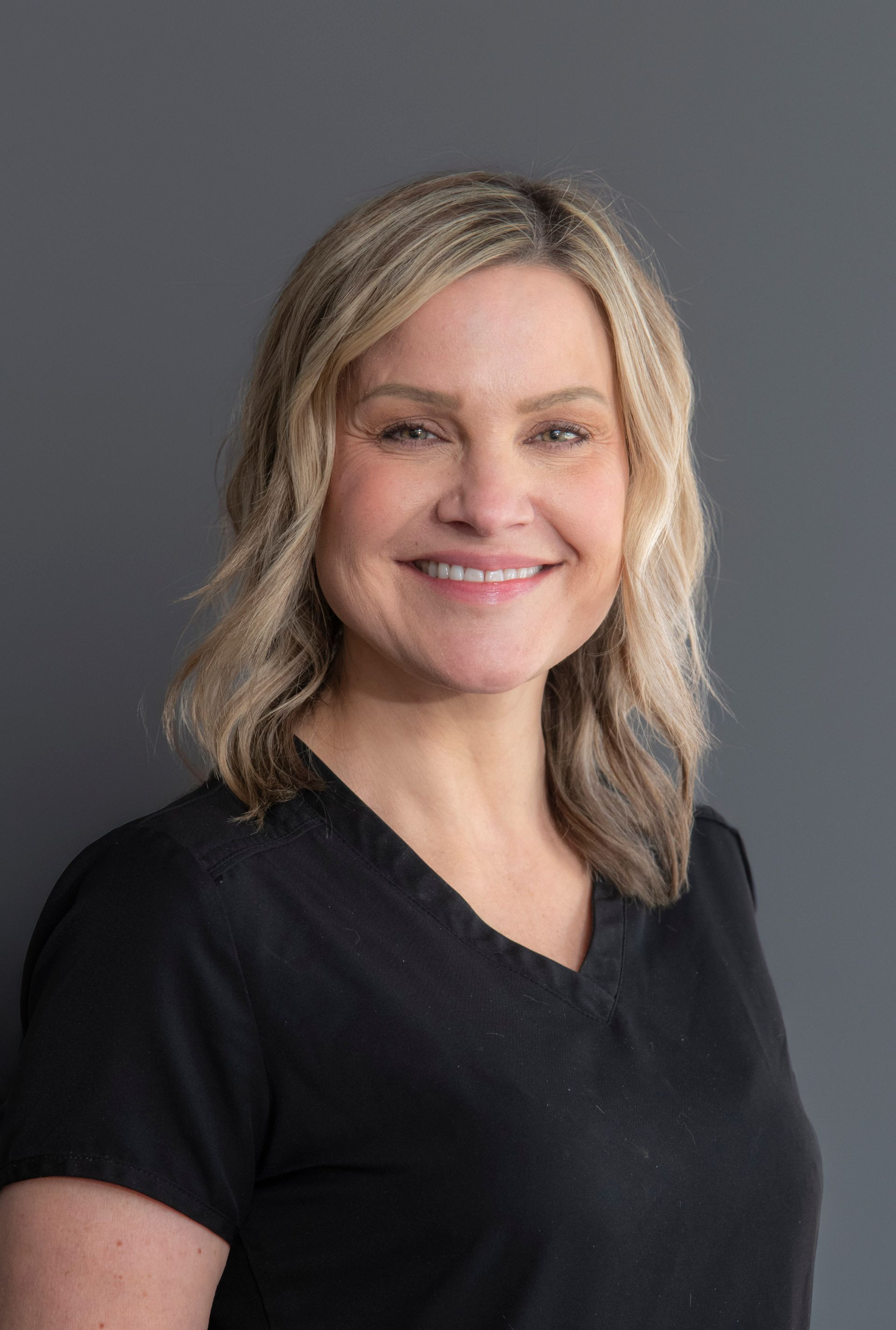 Woman with blonde hair smiling, wearing a black v-neck shirt against a gray background.