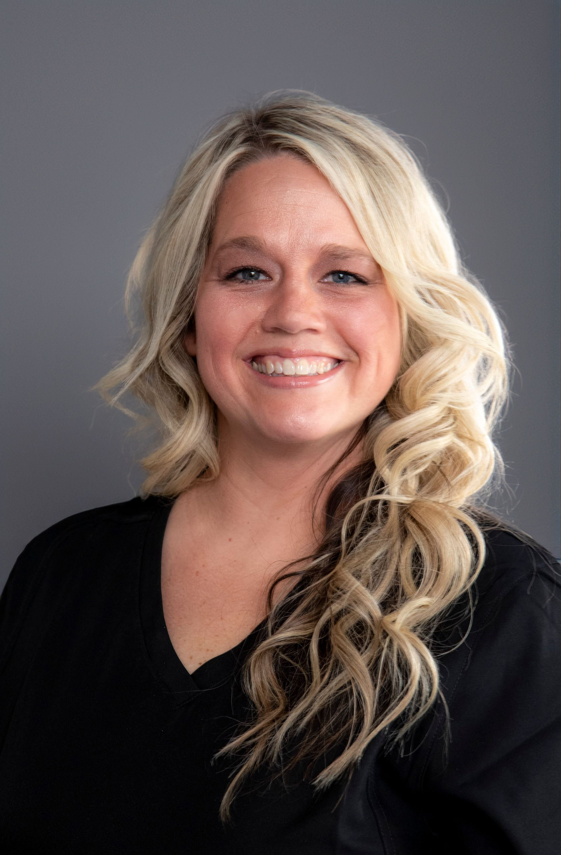Blonde woman smiling at camera, wearing black top, against a grey background.