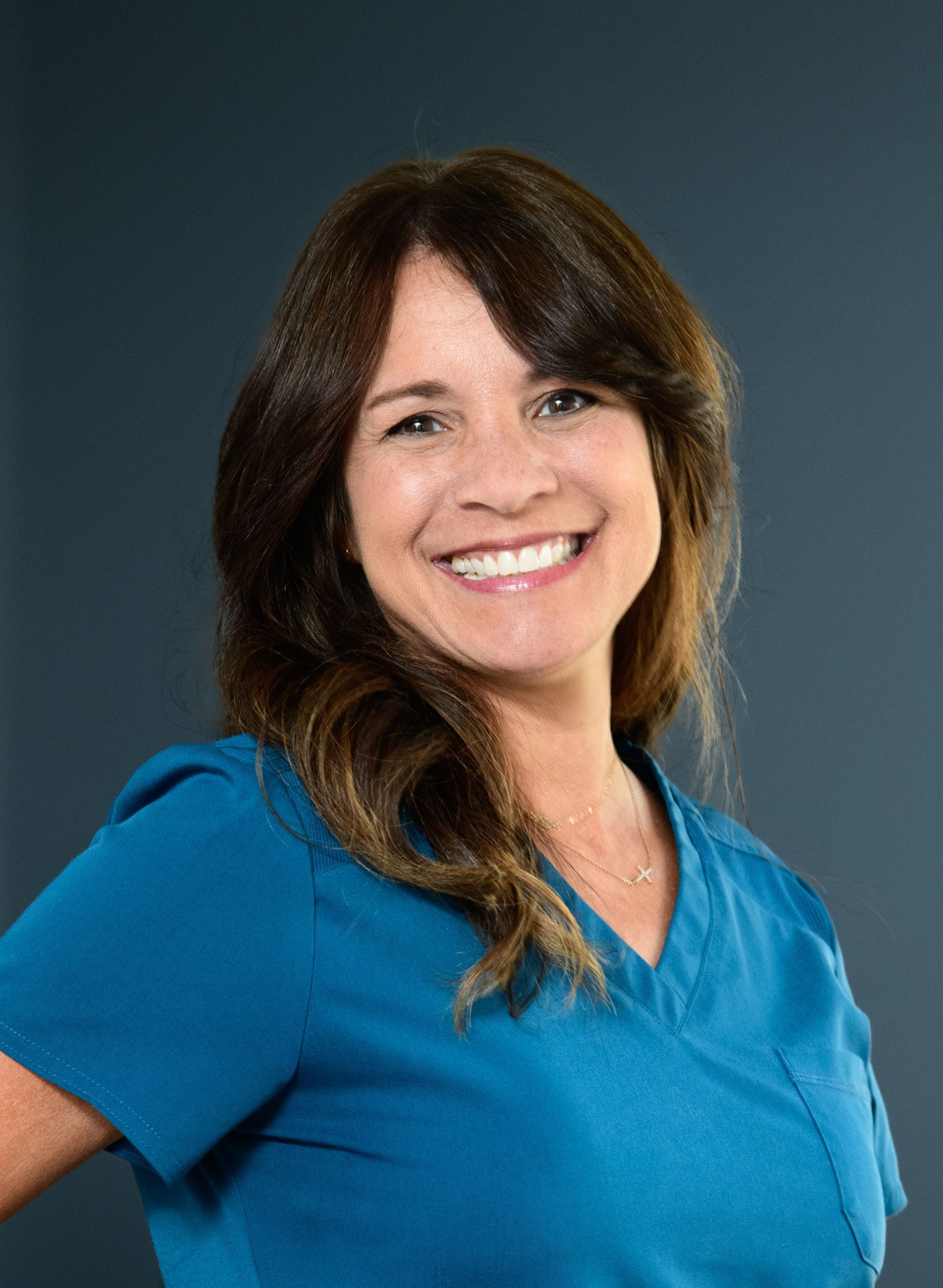 Woman with long brown hair smiles, wearing a teal v-neck top, against a gray background.