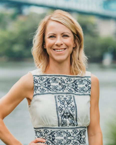 Blonde woman smiling, wearing white dress with blue detail, standing outside near water.