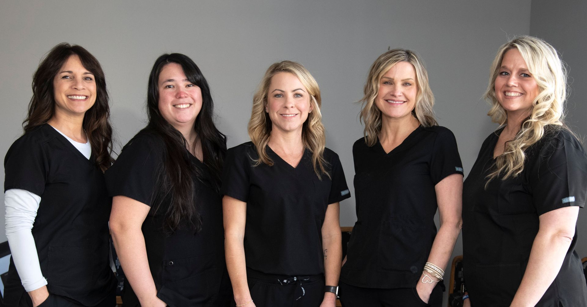 Five women in black scrubs smiling, posed indoors.