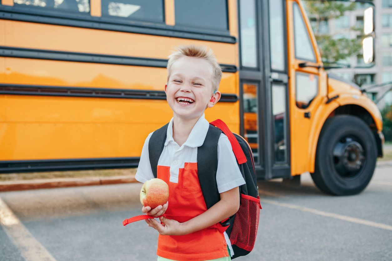 Boy with backpack and apple smiles by a school bus.