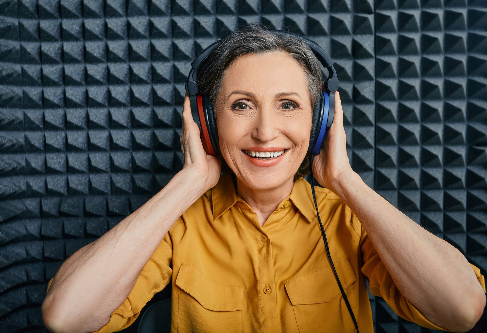 Woman with headphones in soundproof booth, smiling, wearing yellow shirt.