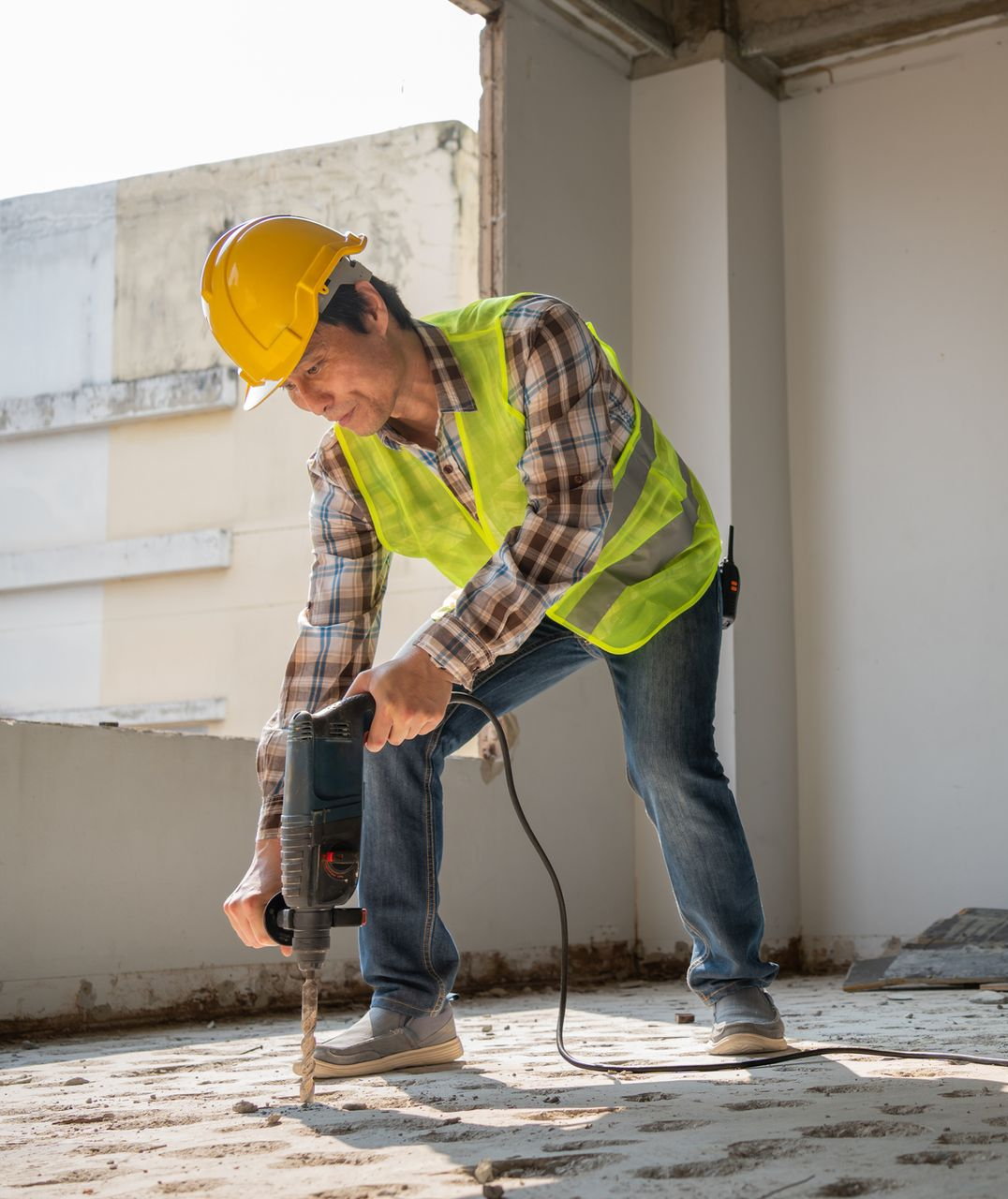 Construction worker drilling concrete floor, wearing yellow hard hat and vest.