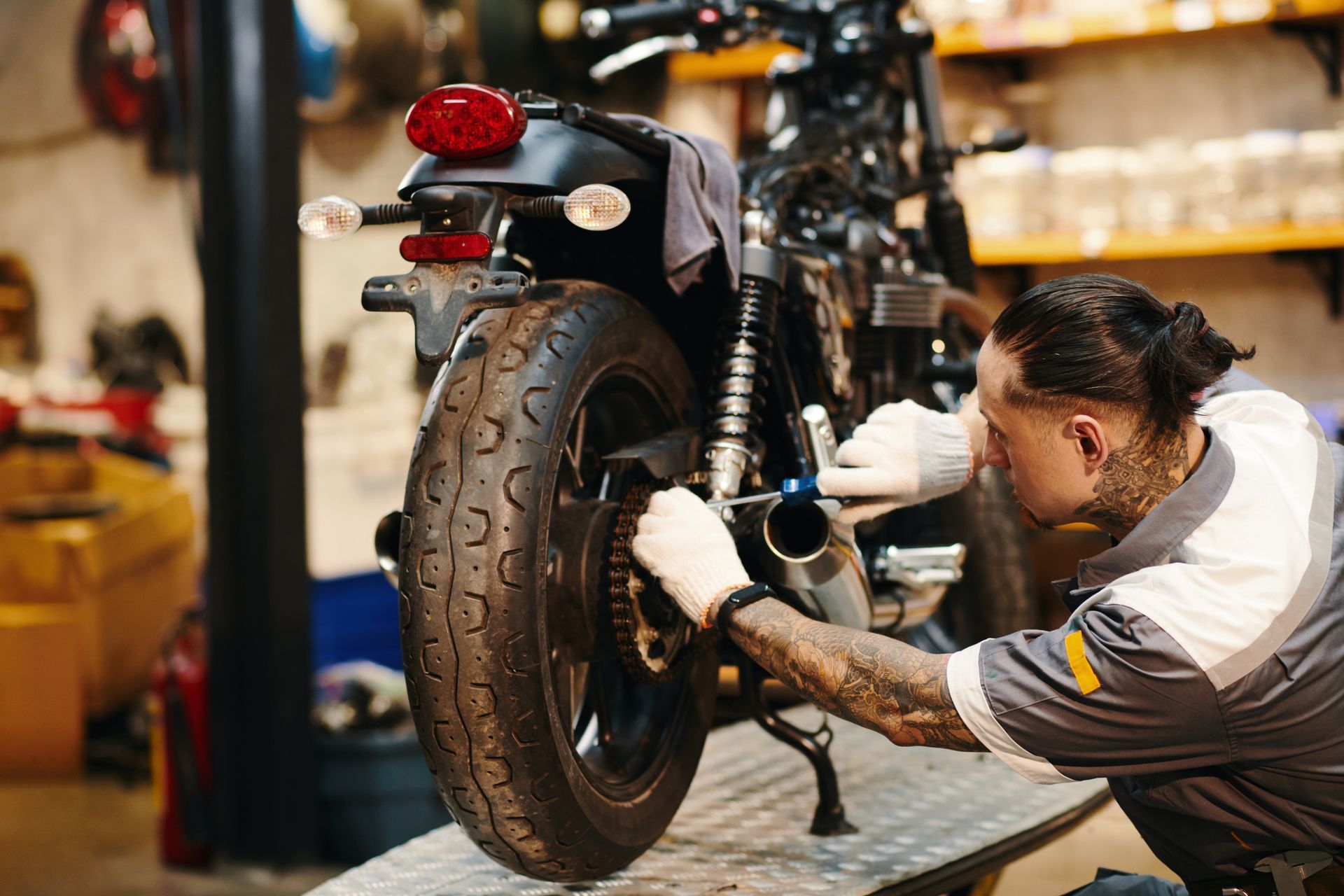 Mechanic working on a motorcycle's rear tire in a garage. He's wearing gloves and has tattoos.