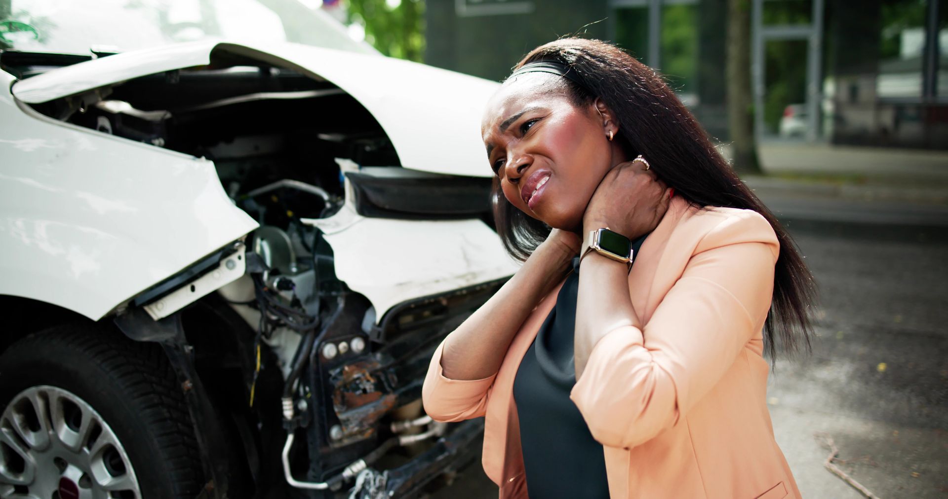 Woman clutches her neck in pain after a car accident; damaged white vehicle in background.